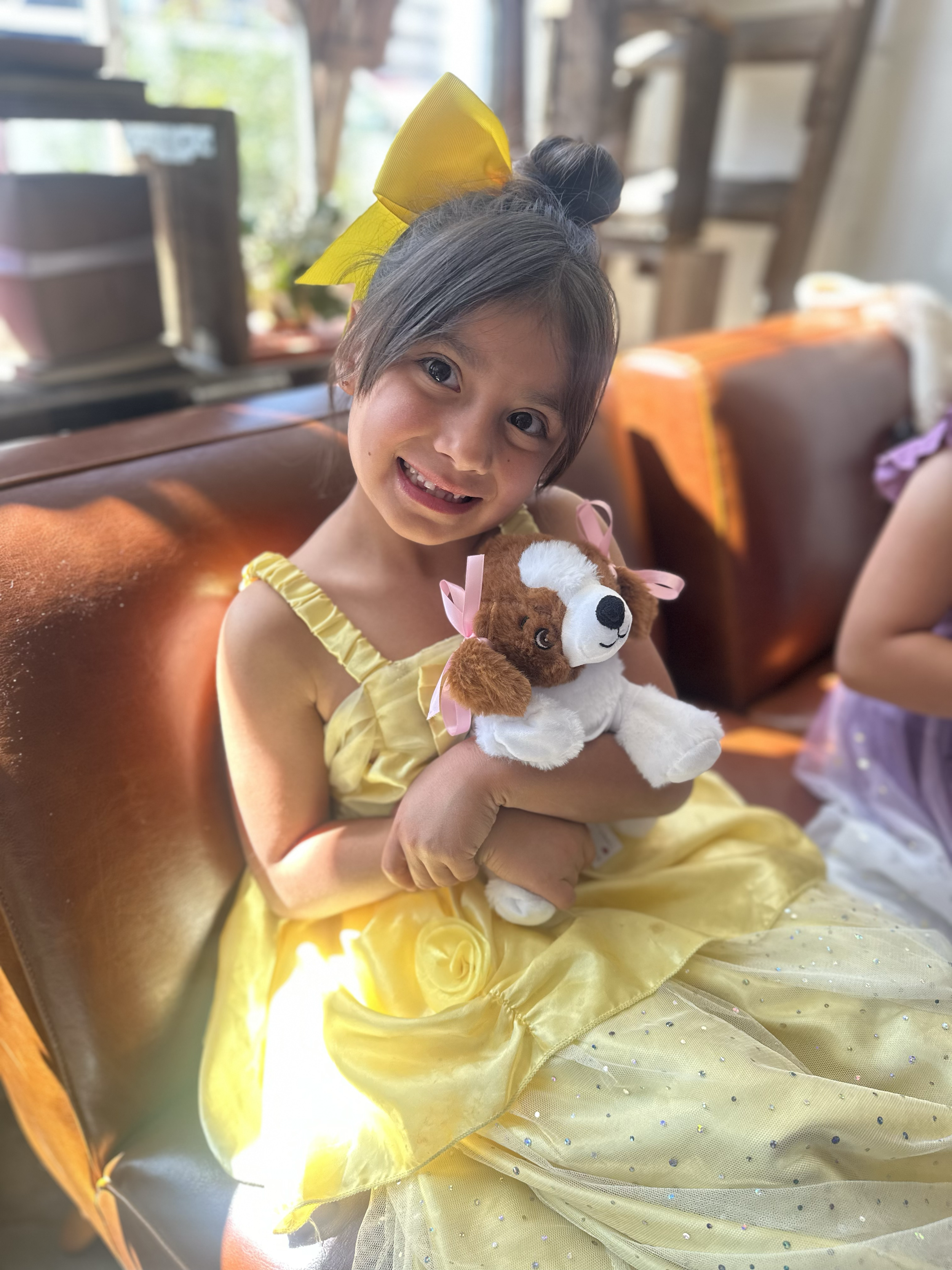 A young girl in a yellow dress sitting on a brown leather couch, smiling and holding a plush dog toy with pink ribbons.