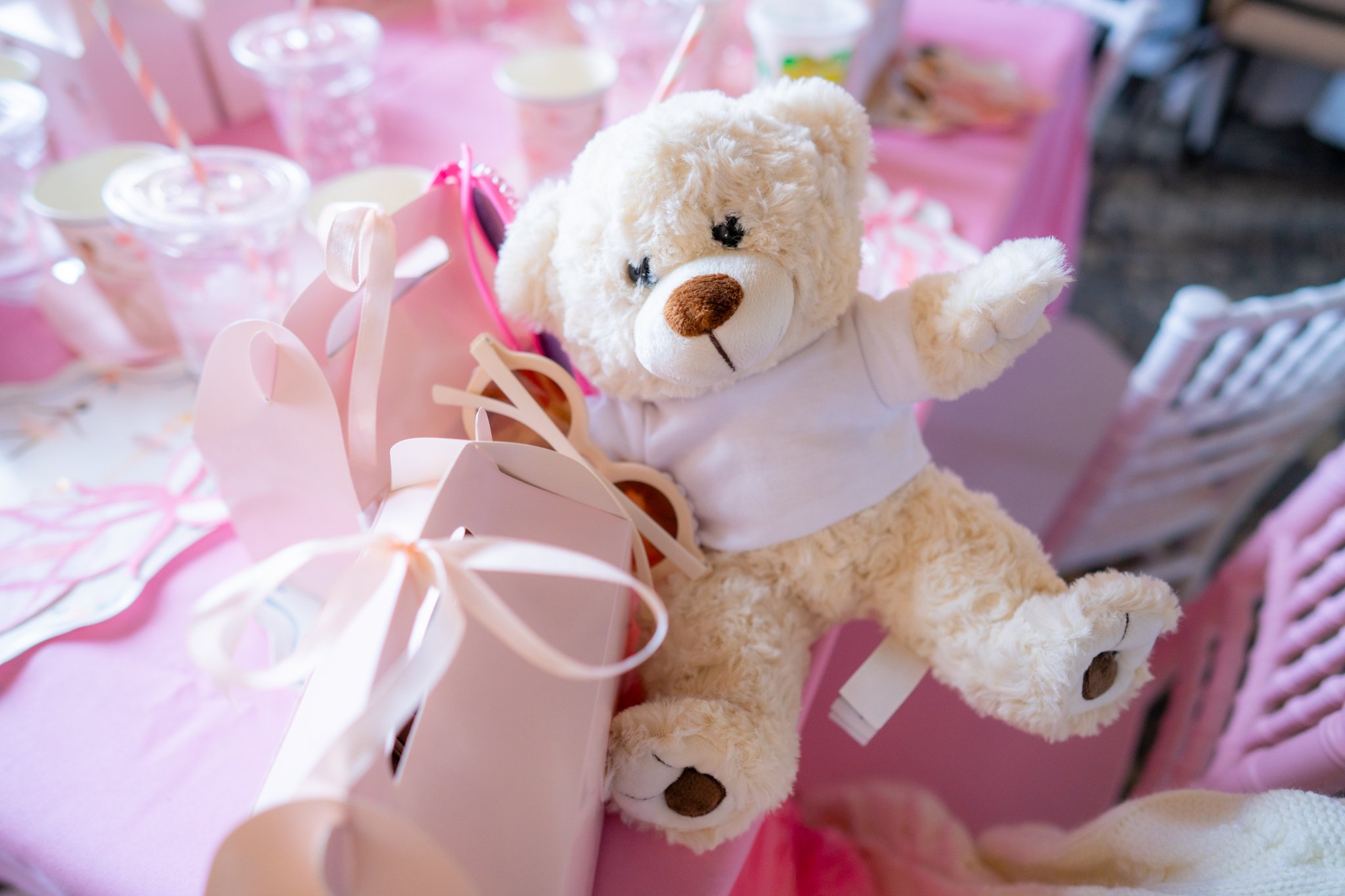 A plush teddy bear wearing a white t-shirt sitting among pink gift boxes and cups on a pink tablecloth, with pink chairs in the background.