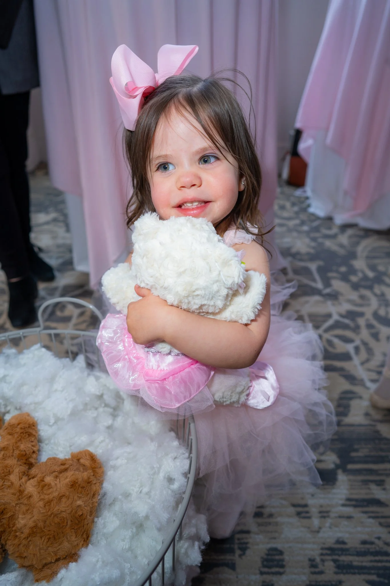 A young girl with brown hair and blue eyes, wearing a pink tutu dress and a large pink bow on her head, hugging a white teddy bear at a decorated event.