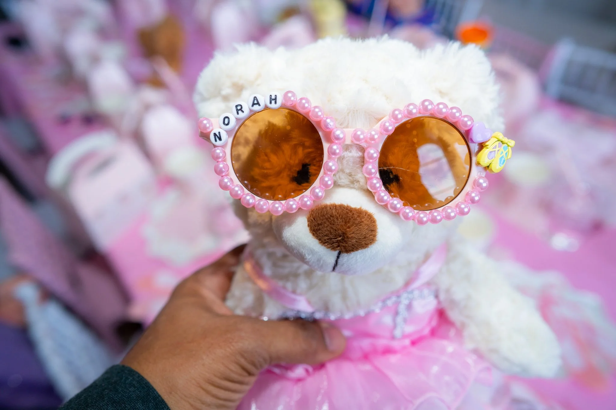 A stuffed teddy bear wearing pink sunglasses decorated with pearl beads that spell out 'NORA H' and a pink heart and butterfly charm, dressed in a pink dress, held in a person's hand with a blurred background of pink items.