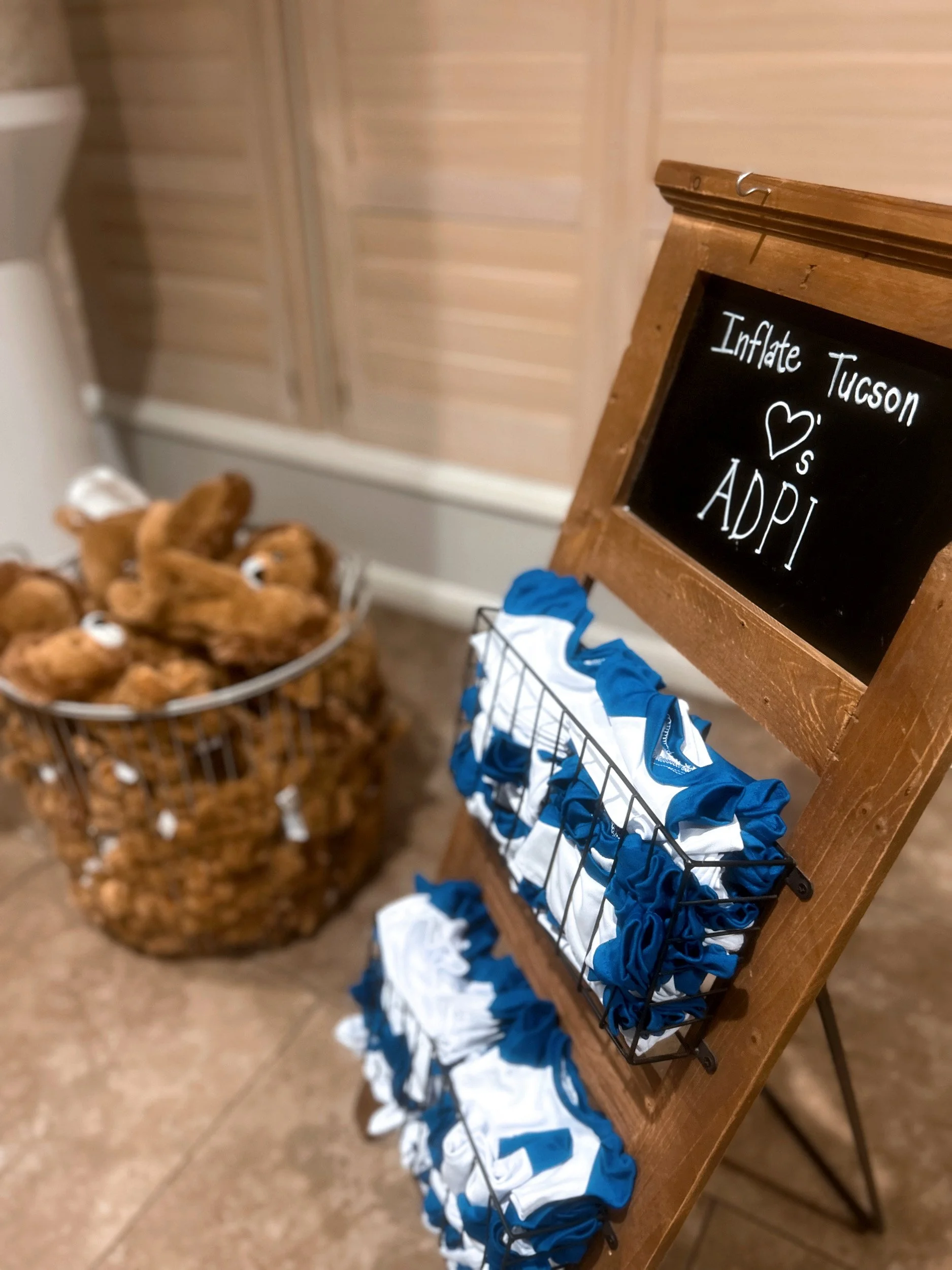 Towels in blue and white colors folded on a black rack next to a sign that says 'Inflate Tucson ❤️s ADPI', with a basket of teddy bears in the background.