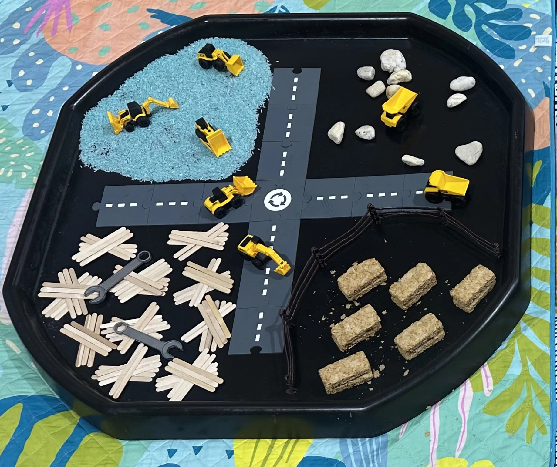A game board with toy construction vehicles, rocks, wooden sticks, cereal bars, and paper road sections, resembling a construction site simulation.