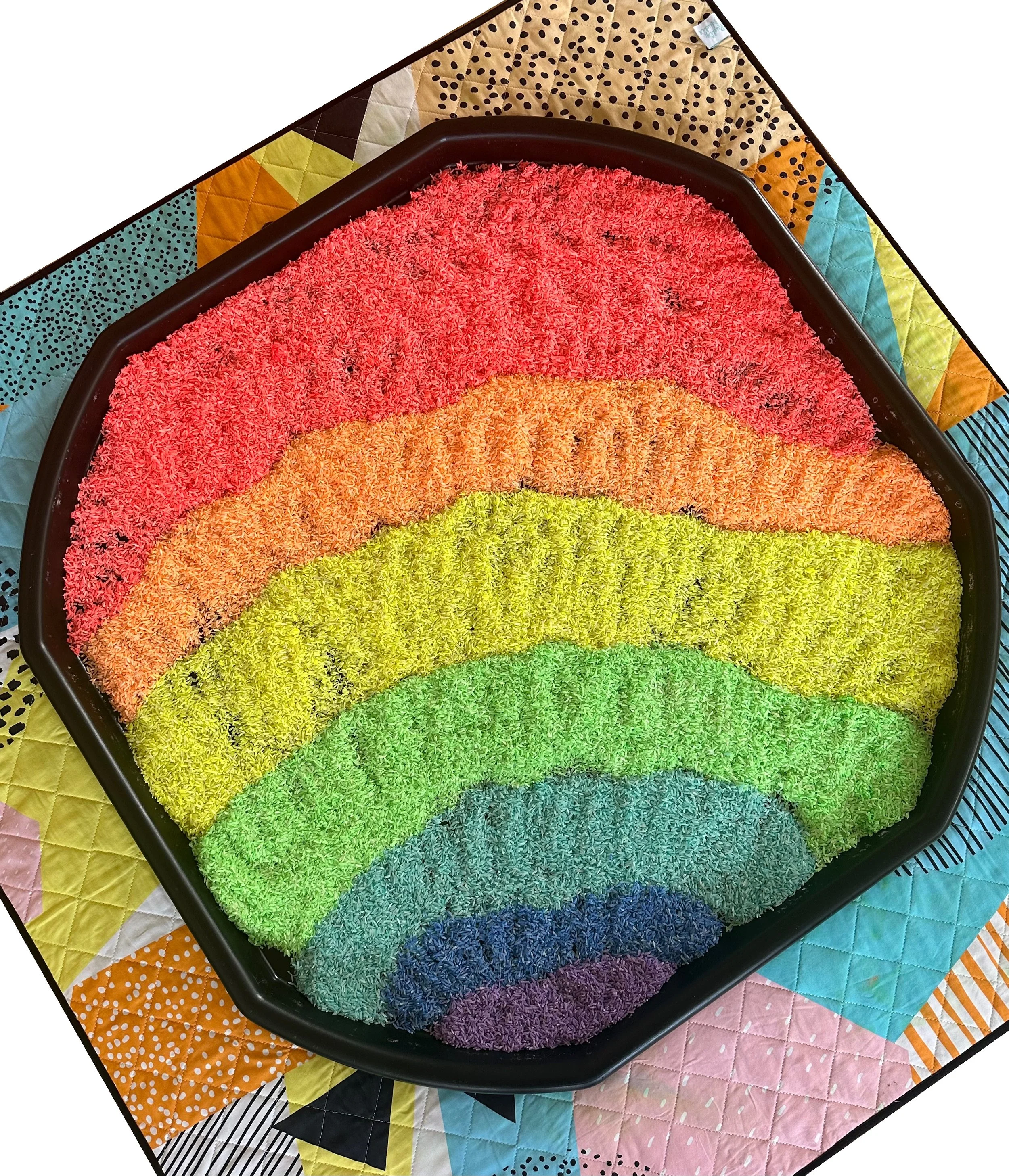 Colorful rainbow-shaped textured rug with multiple colors including pink, orange, yellow, green, blue, and purple, on top of a multicolored patchwork quilt.