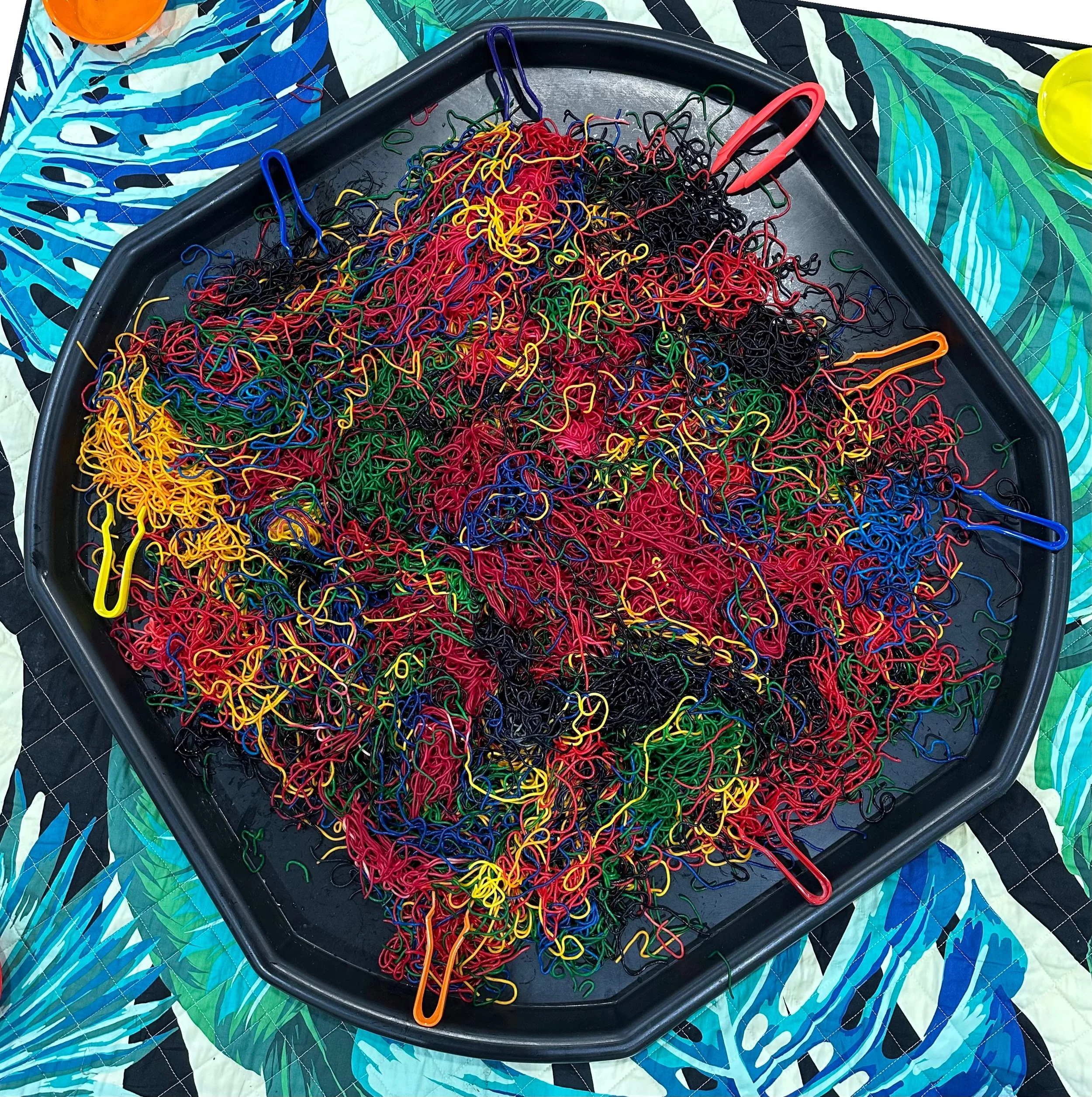 A large black tray filled with colorful rubber bands and paperclips, placed on a patterned fabric surface.