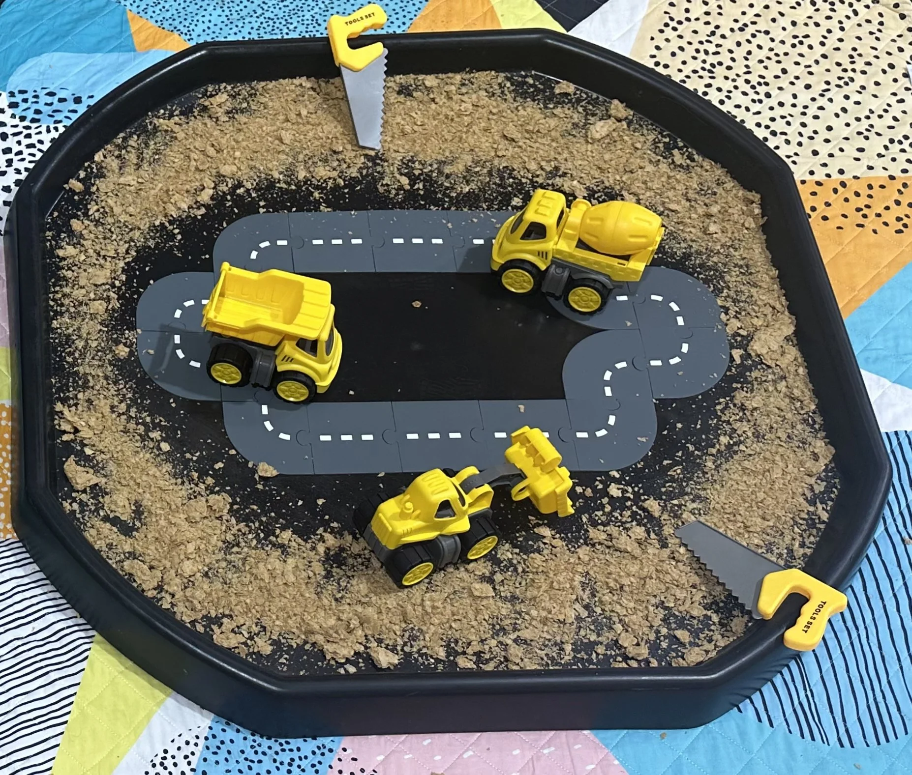 A children's sandbox with a black border, a gray winding road patterned mat, and three yellow toy construction vehicles. The sandbox is filled with sand and has four small yellow saws labeled 'TOOLS SET' in the corners.