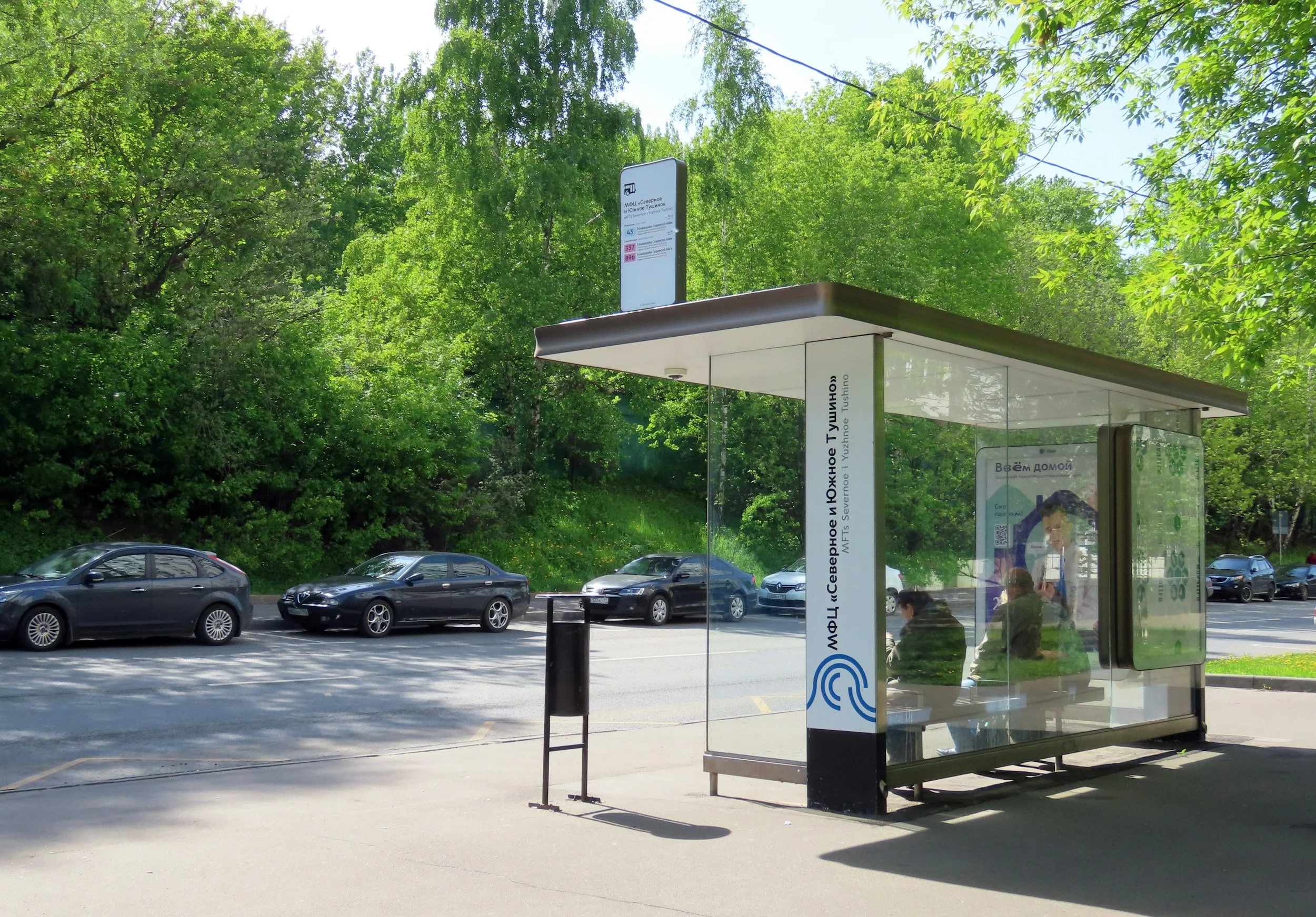 Bus stop shelter with two people seated inside, along a street lined with parked cars and green trees in the background.