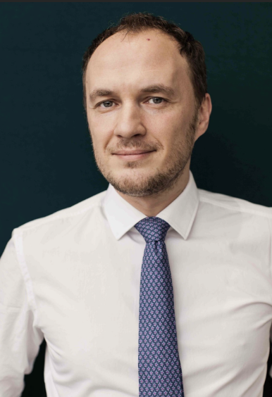 A man in a white shirt and patterned tie standing against a dark background.
