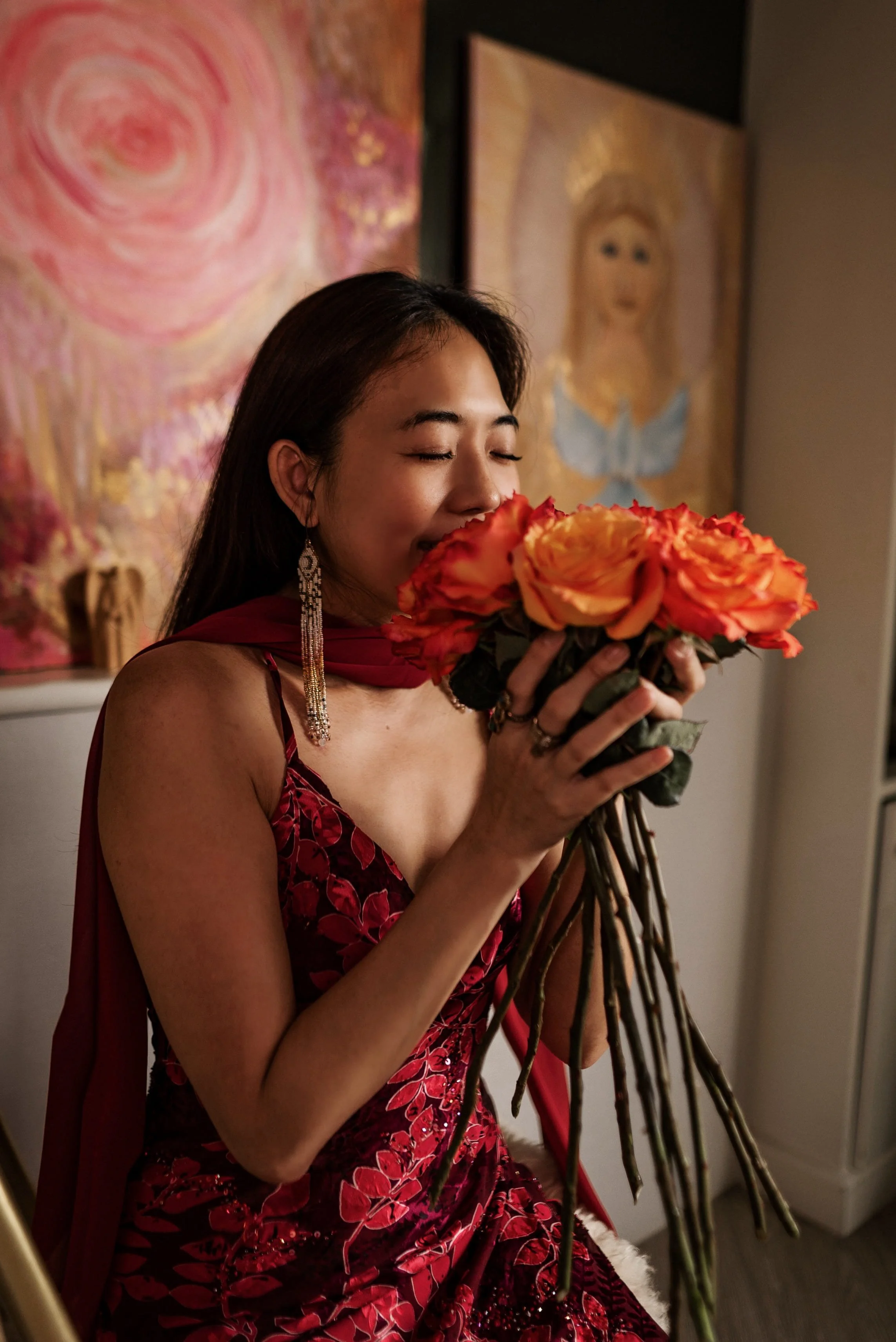 a dark-haired woman in a red dress smelling orange roses, illustrating her 1:1 womb blessing session in Bangkok Thailand.