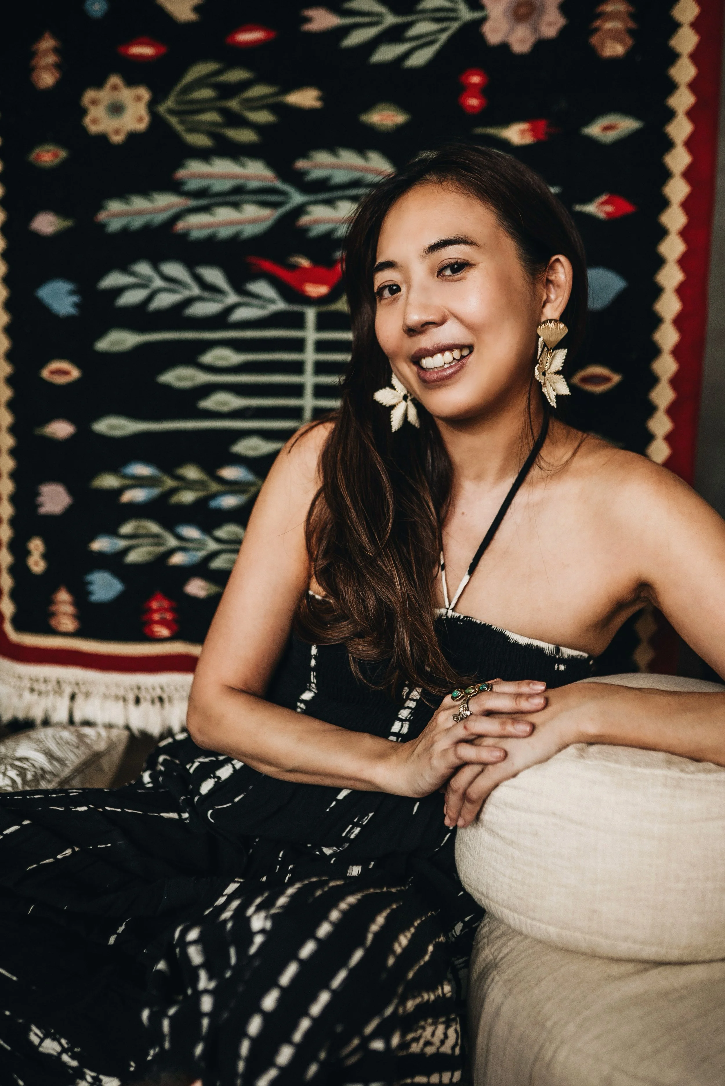 A portrait of a dark-haired woman waring a black dress and statement earrings, smiling to the camera