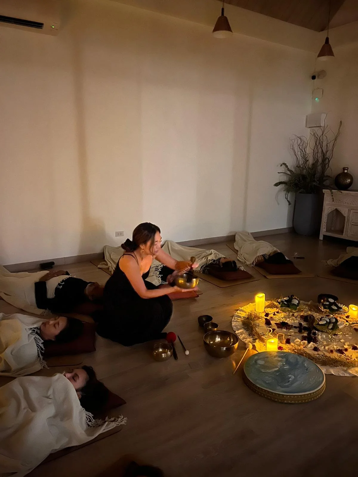 A group sound healing session in Bangkok where the practitioner is in the middle in front of an altar playing. a crystal bowl while participants are laying down in a circle on yoga mats