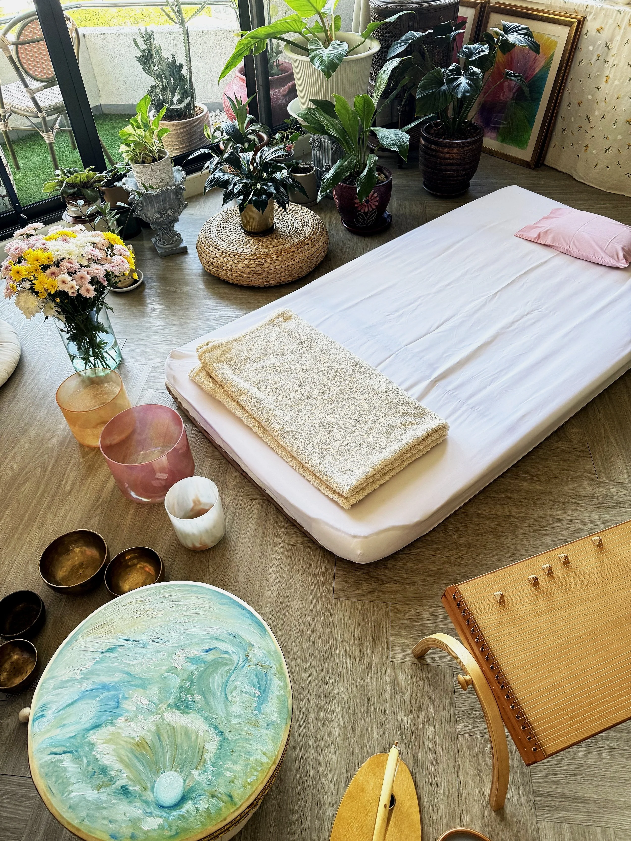 Image of a sacred space, mattress surrounded by flowers, plants, crystal singing bowls, and music instruments, illustrating a 1:1 sound healing session in bangkok thailand