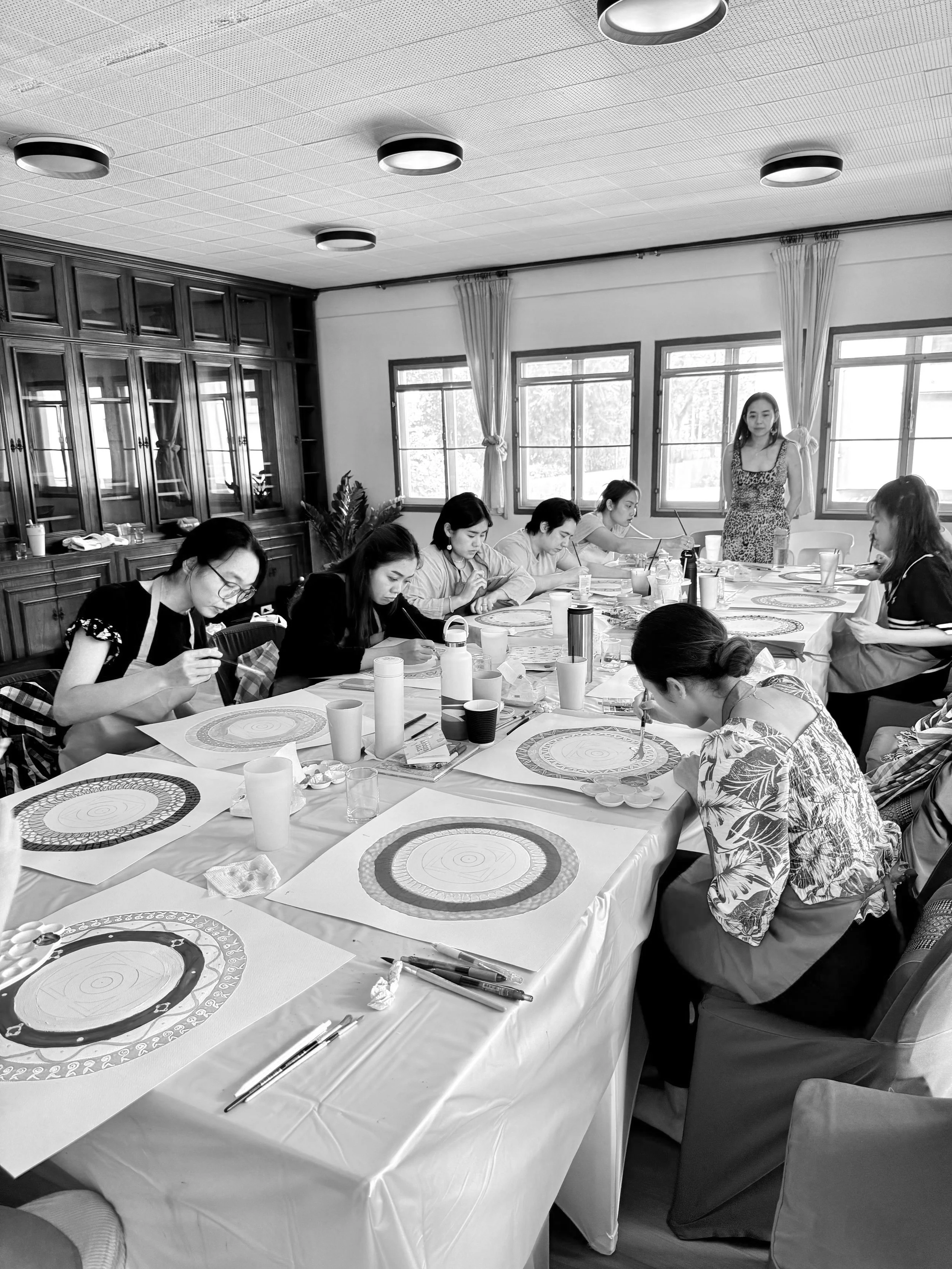 black and white photo of a group sitting behind a table, drawing mandalas whilst the teacher is observing them
