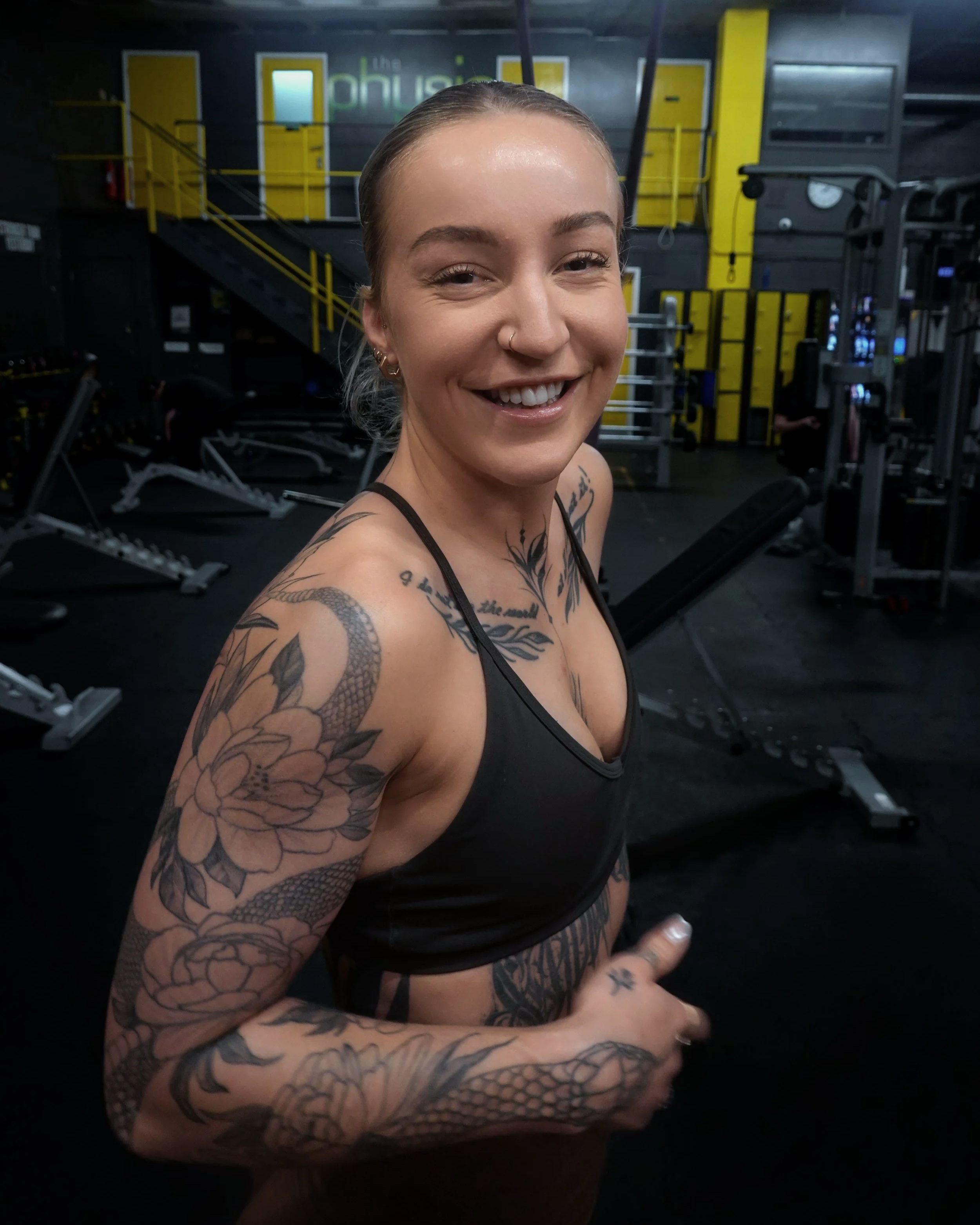 A woman smiling at the camera inside a gym, with tattoos on her arms and a nose ring, wearing a black sports bra.