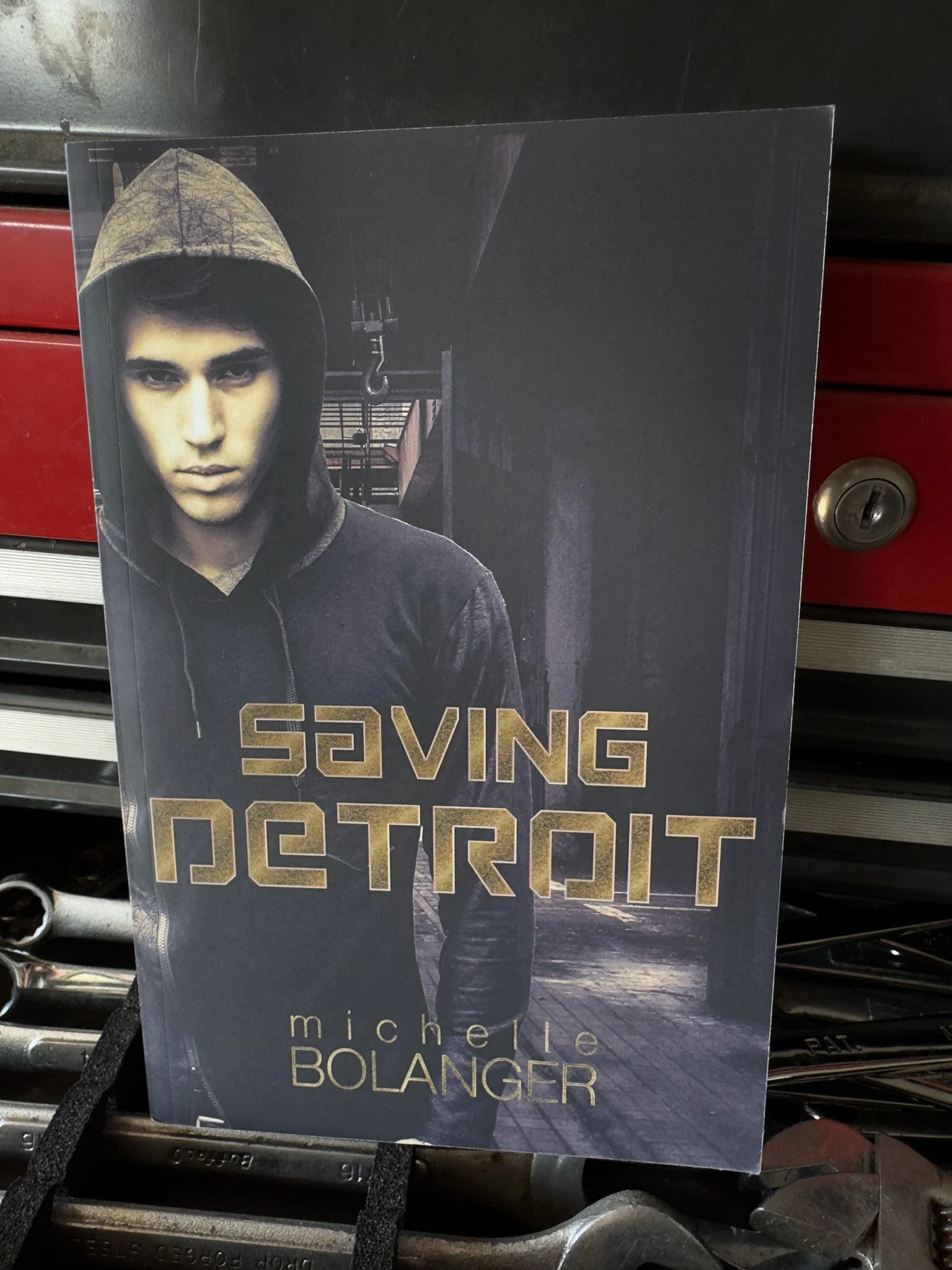 Book cover featuring a young man with dark hair and a hoodie, with a cityscape background. The title includes the words 'Saving Detroit' and the author's name Michele Bolanger.