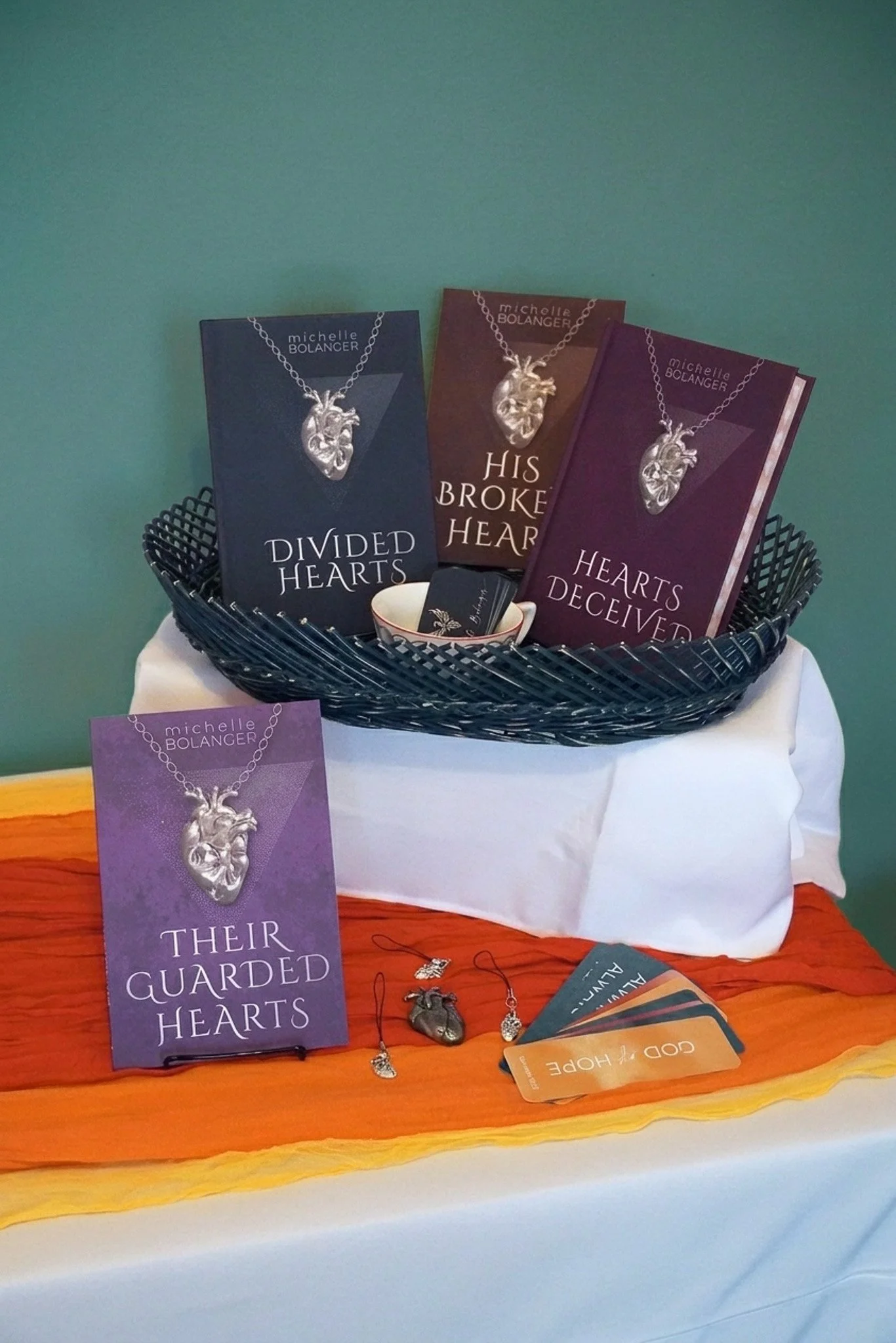 Books in a basket and one on the table next to a metal heart necklace and bookmarks with a teacup filled with business cards