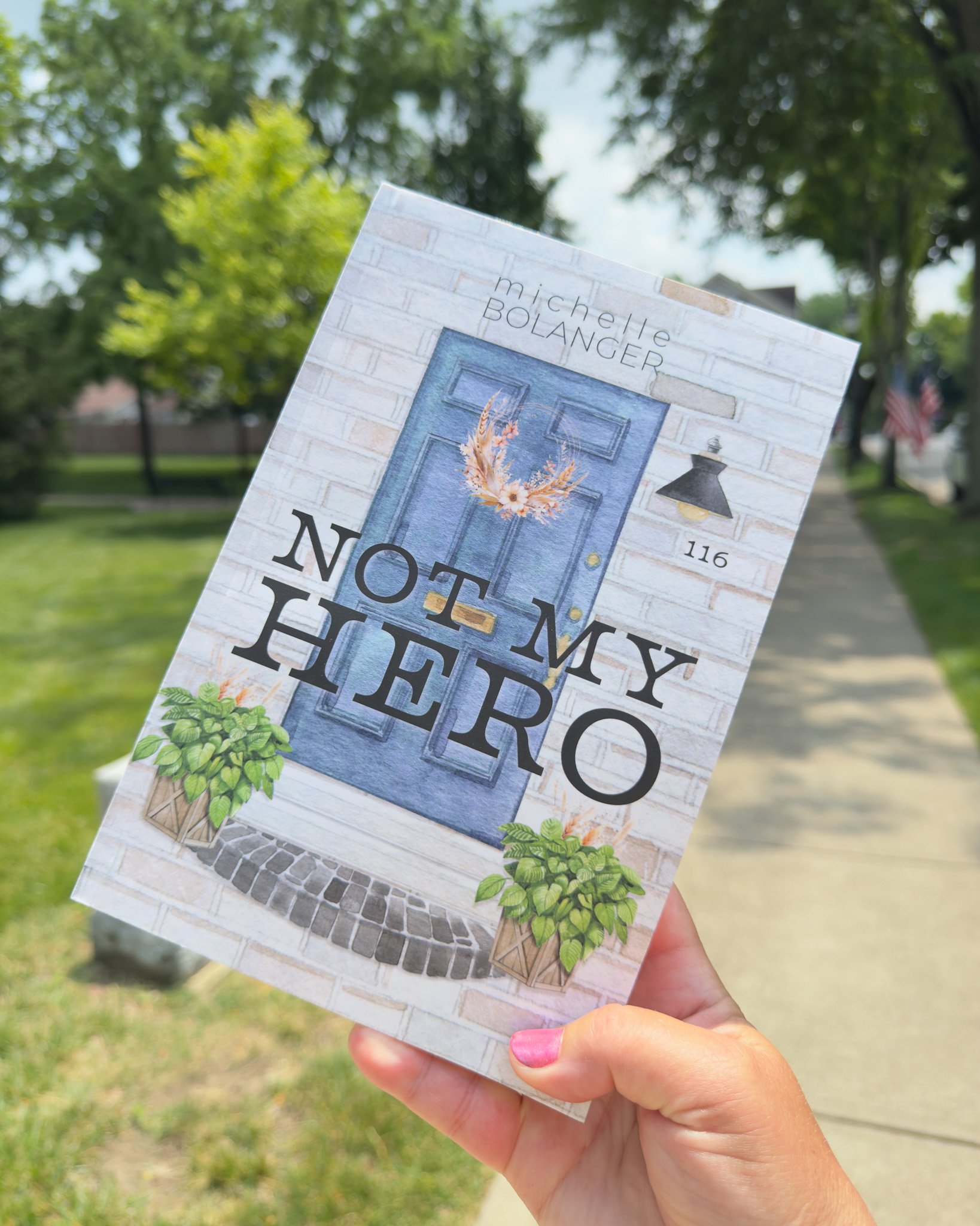 A person holding an election flyer in front of a park with trees, a sidewalk, and American flags, with pink nail polish on their fingernails.
