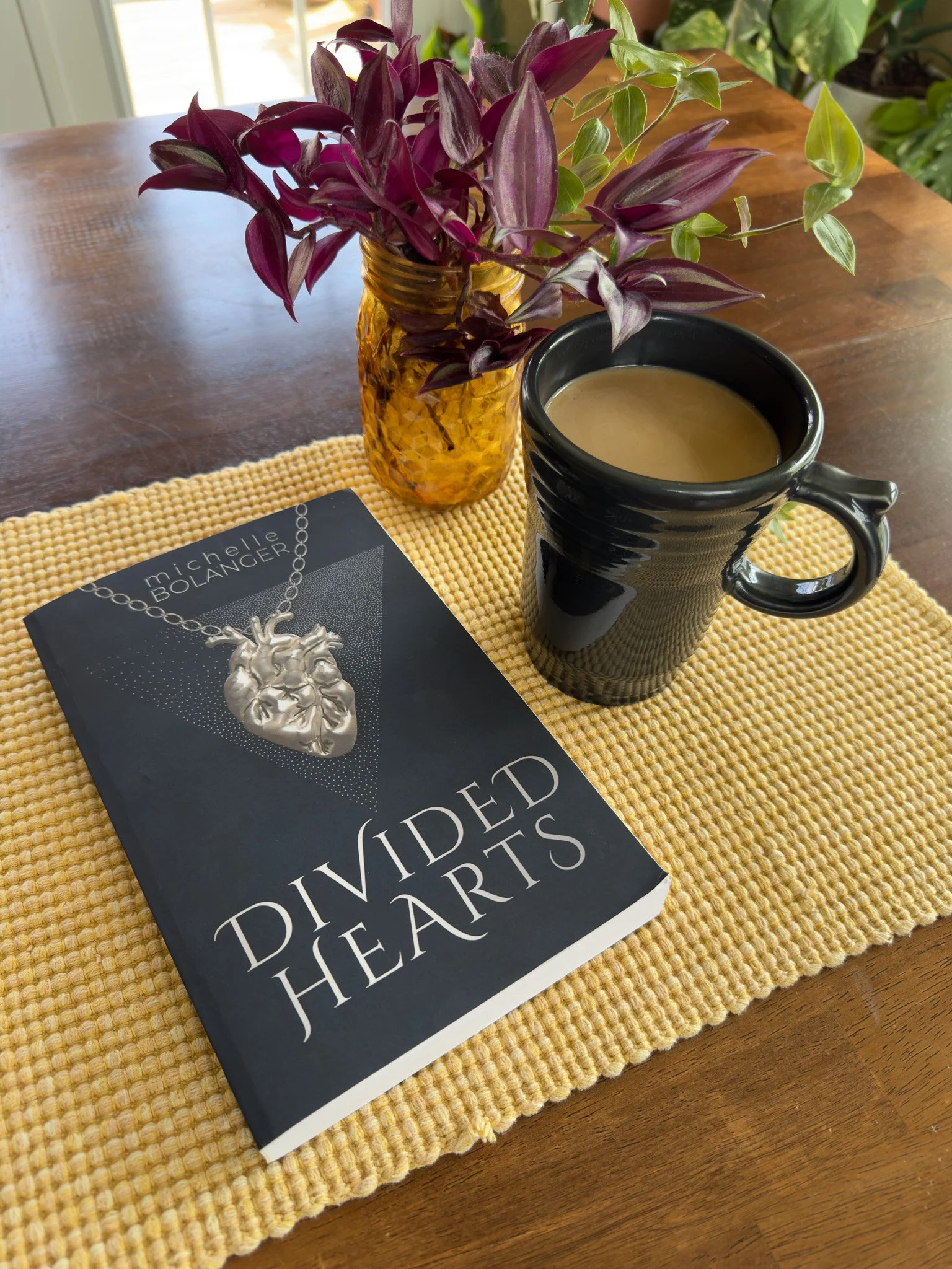 A black mug filled with coffee, a yellow textured glass vase with purple and green plants, a book titled "Divided Hearts" by Michelle Bolanger, all on a yellow woven placemat on a wooden table.