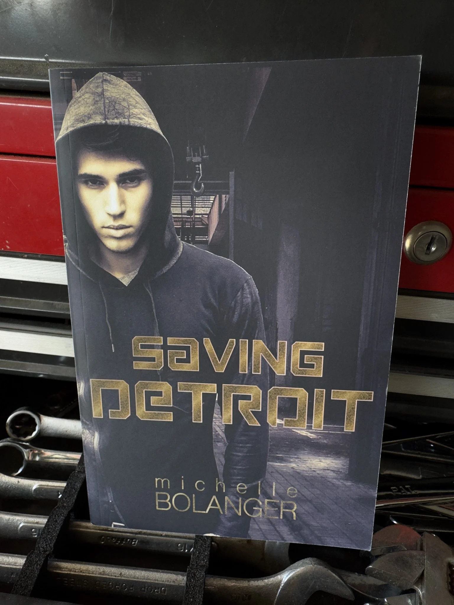 A book cover featuring a young man with dark hair wearing a hoodie with a camouflage hood, set against an urban background with shadows and a crane hook. The title is "Saving Detroit" and the author is Michelle Bolanger.