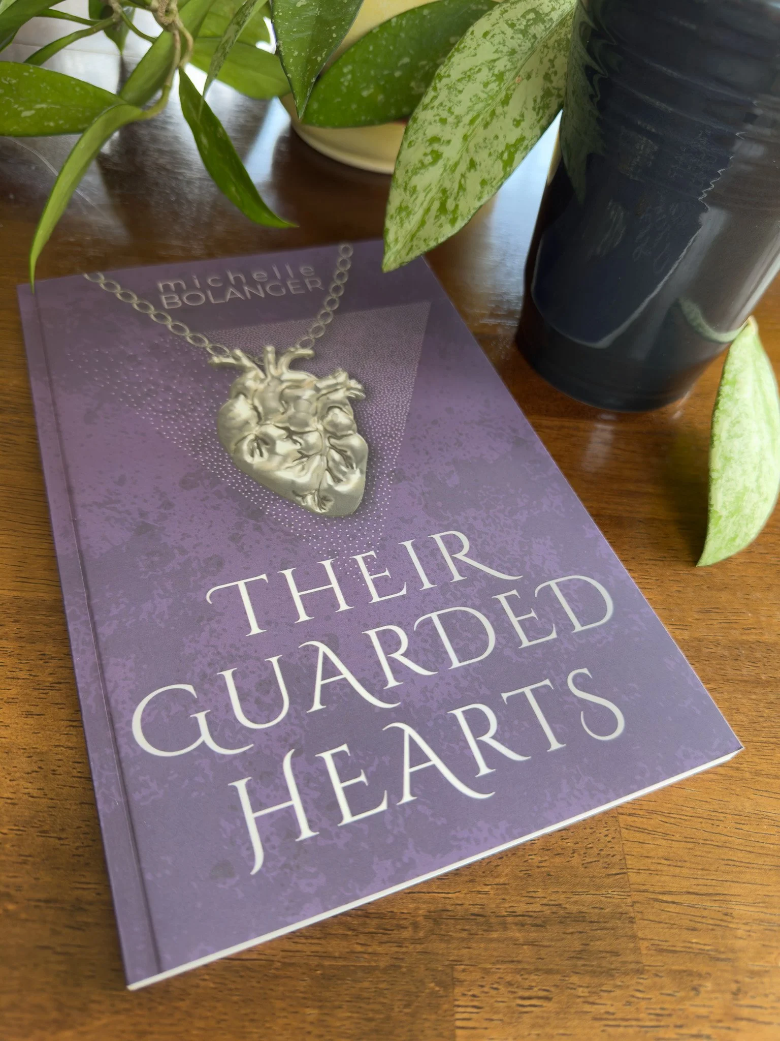 A book titled 'Their Hidden Guardians' placed on a wooden surface, partially covered by a green potted plant, a black vase, and a silver heart-shaped jewelry piece resting on purple paper.