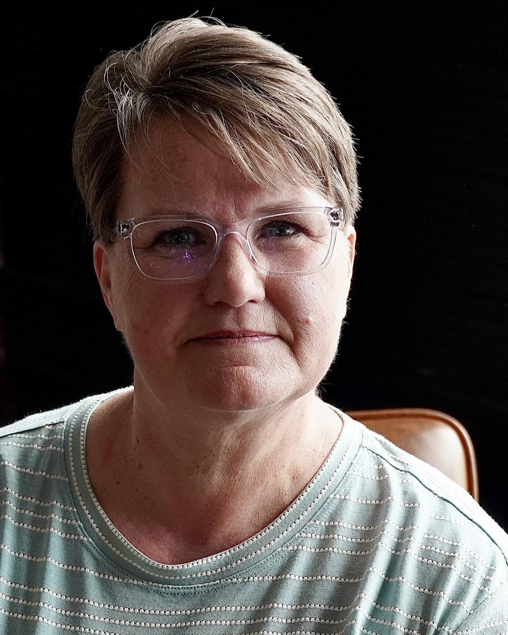 Close-up of a woman's face, wearing glasses, with short light-colored hair, smiling slightly, against a black background, and dressed in a light-colored striped shirt.