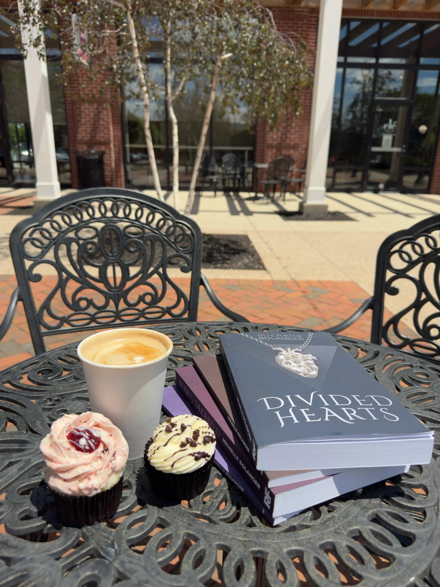 Outdoor patio table with a cup of coffee, three cupcakes, and three books, with a brick and glass building in the background.