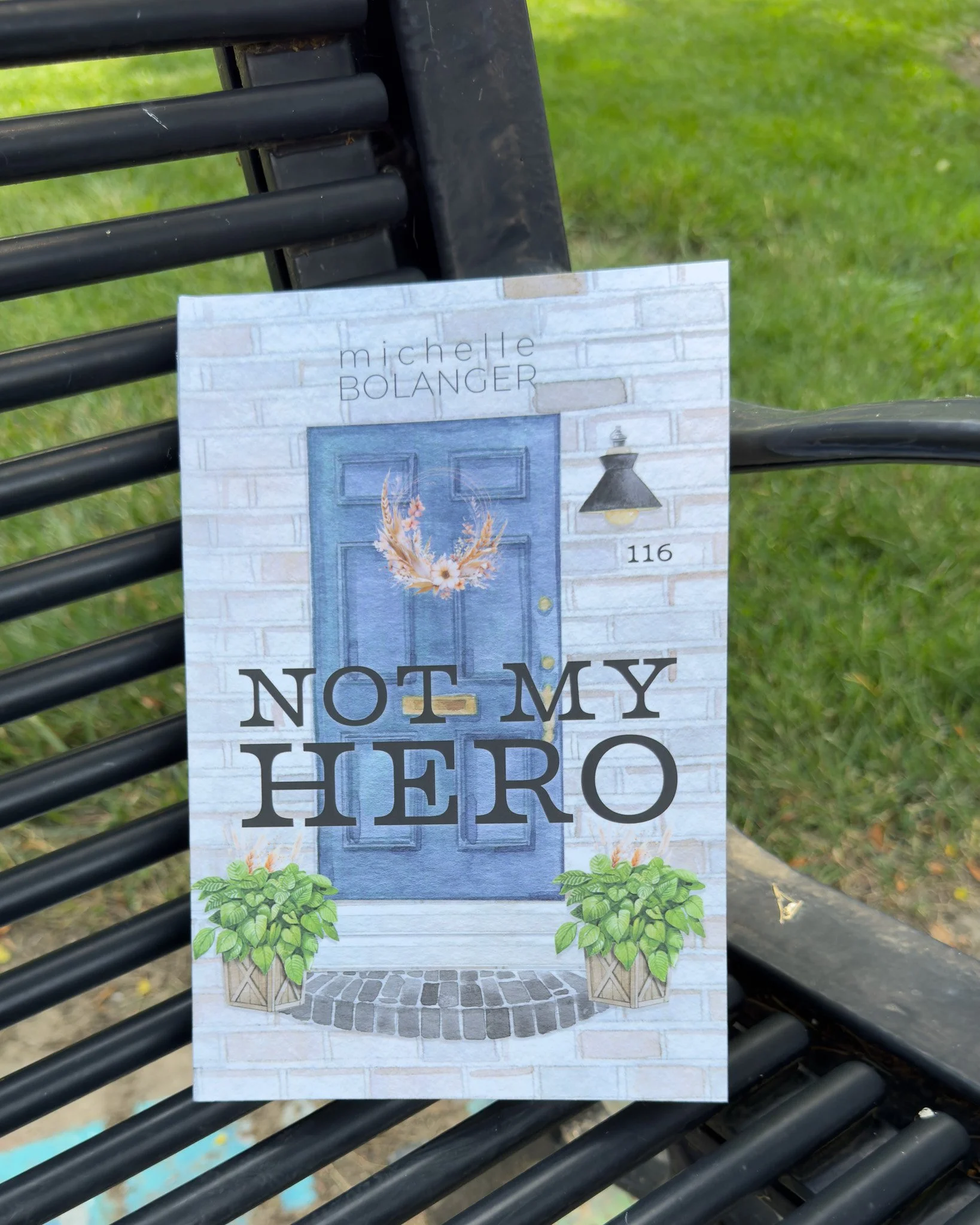 A sign hanging on a black metal bench. The sign features a blue door, plants, and the text 'NOT MY' at the bottom. The background is outdoor grass and a walkway.