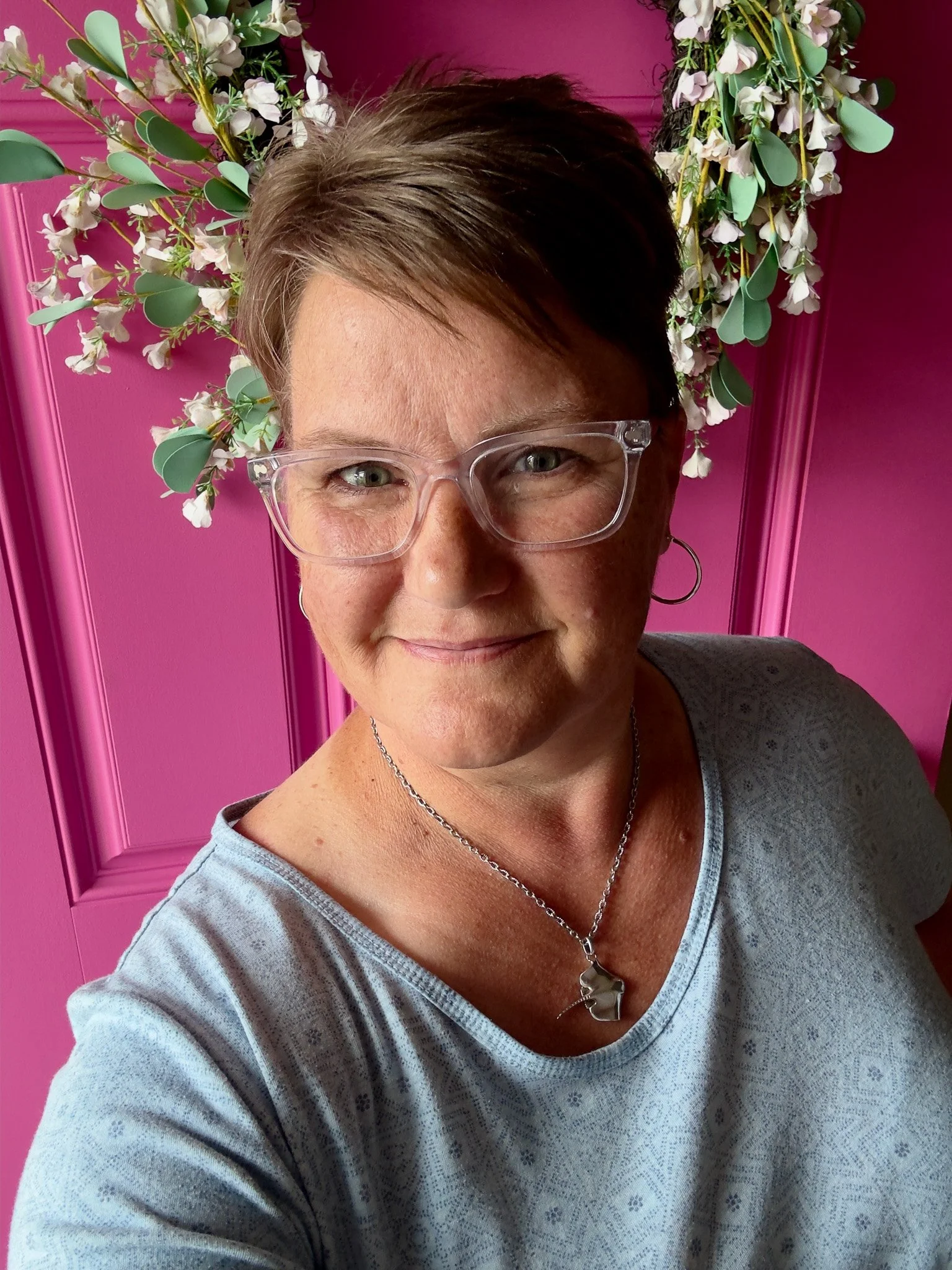 woman wearing glasses and smiling in front of pink door with floral wreath behind her