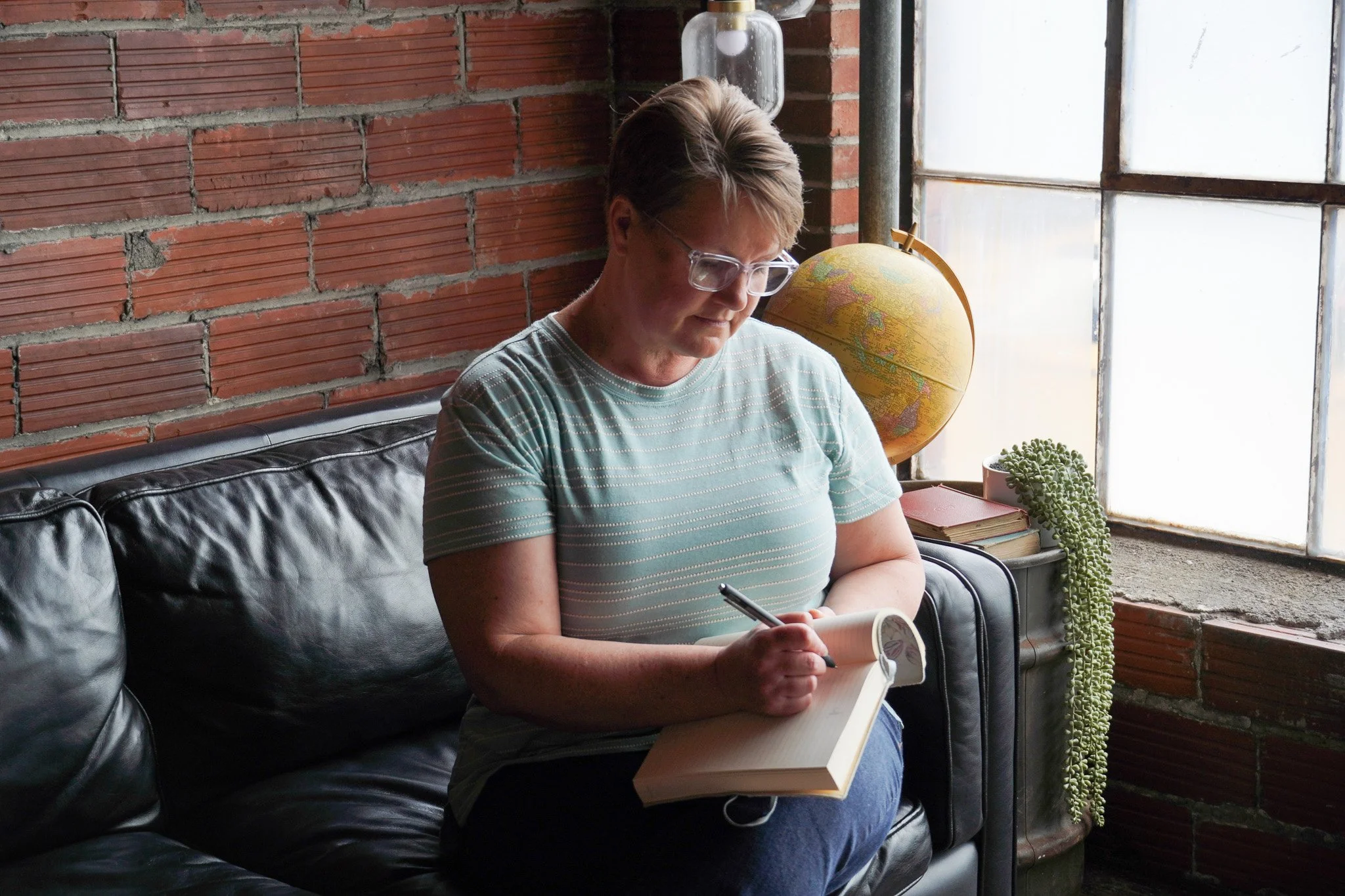 A woman with short hair and glasses sitting on a black leather couch, writing in a notebook with a pen, in front of a brick wall with large windows.