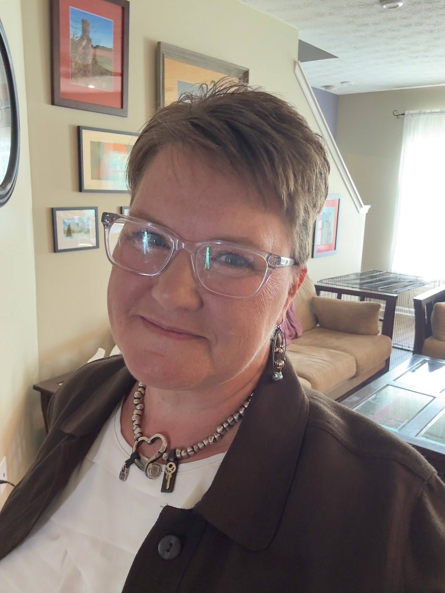 A woman with short brown hair and glasses smiling at the camera, wearing a black jacket, a white shirt, and a necklace with heart-shaped and other charms. The background shows a living room with framed pictures on the wall, a window with curtains, a couch, and a glass-top table.