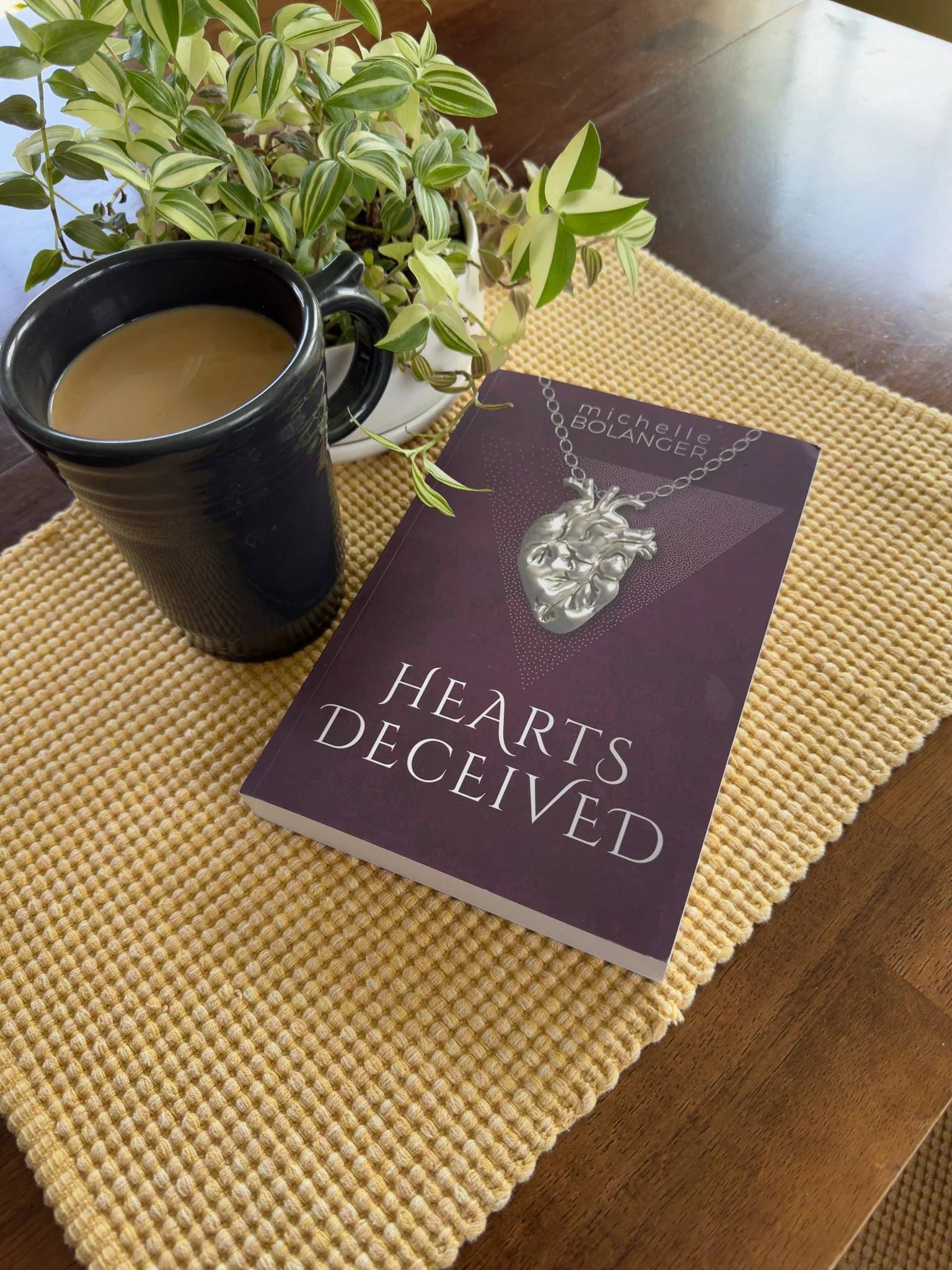 A black coffee mug filled with coffee on a woven yellow placemat, next to a green potted plant and a book titled "Hearts Decieved" by Michelle Bolanger on a wooden table.