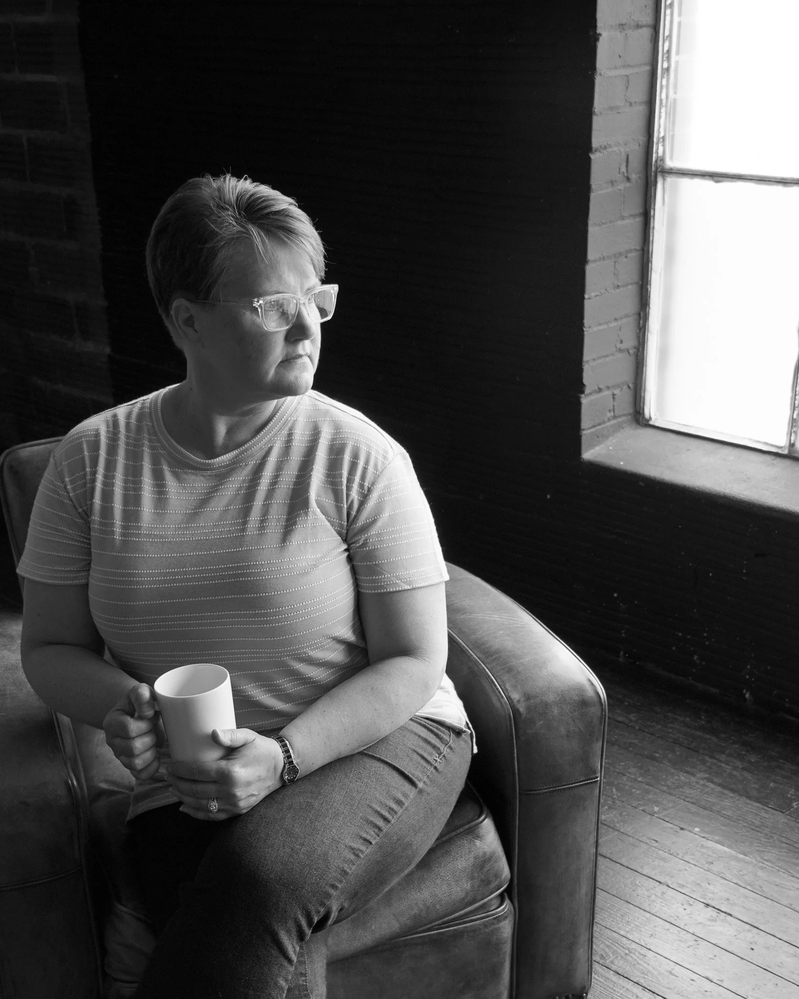 A woman with short hair and glasses sitting on a vintage chair, holding a mug with both hands. She is wearing a striped t-shirt, jeans, and a watch. The setting has a dark wall and a large window with light coming through.