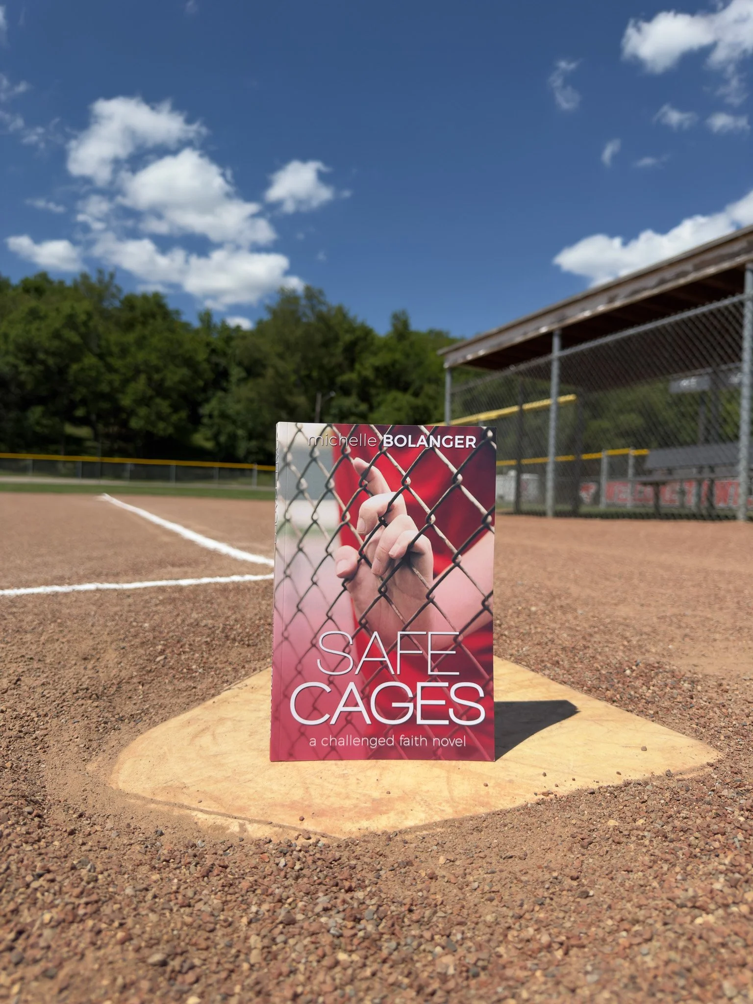 A baseball field with a sign in front of a chain-link fence. The sign reads "Safe Cages, a challenged faith novel" and features an image of a hand gripping the fence.