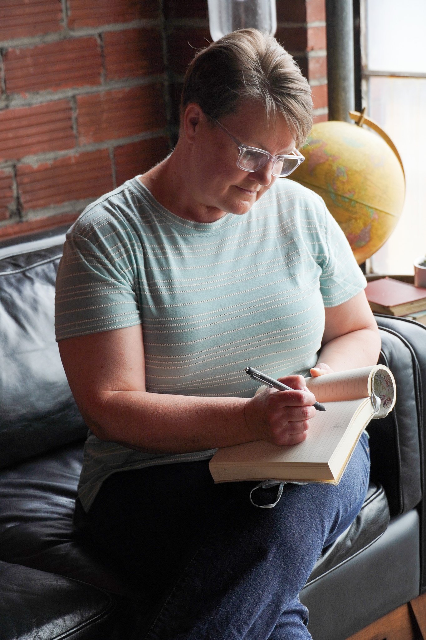 Woman wearing glasses, sitting on a black leather couch writing in a journal in front of a brick wall