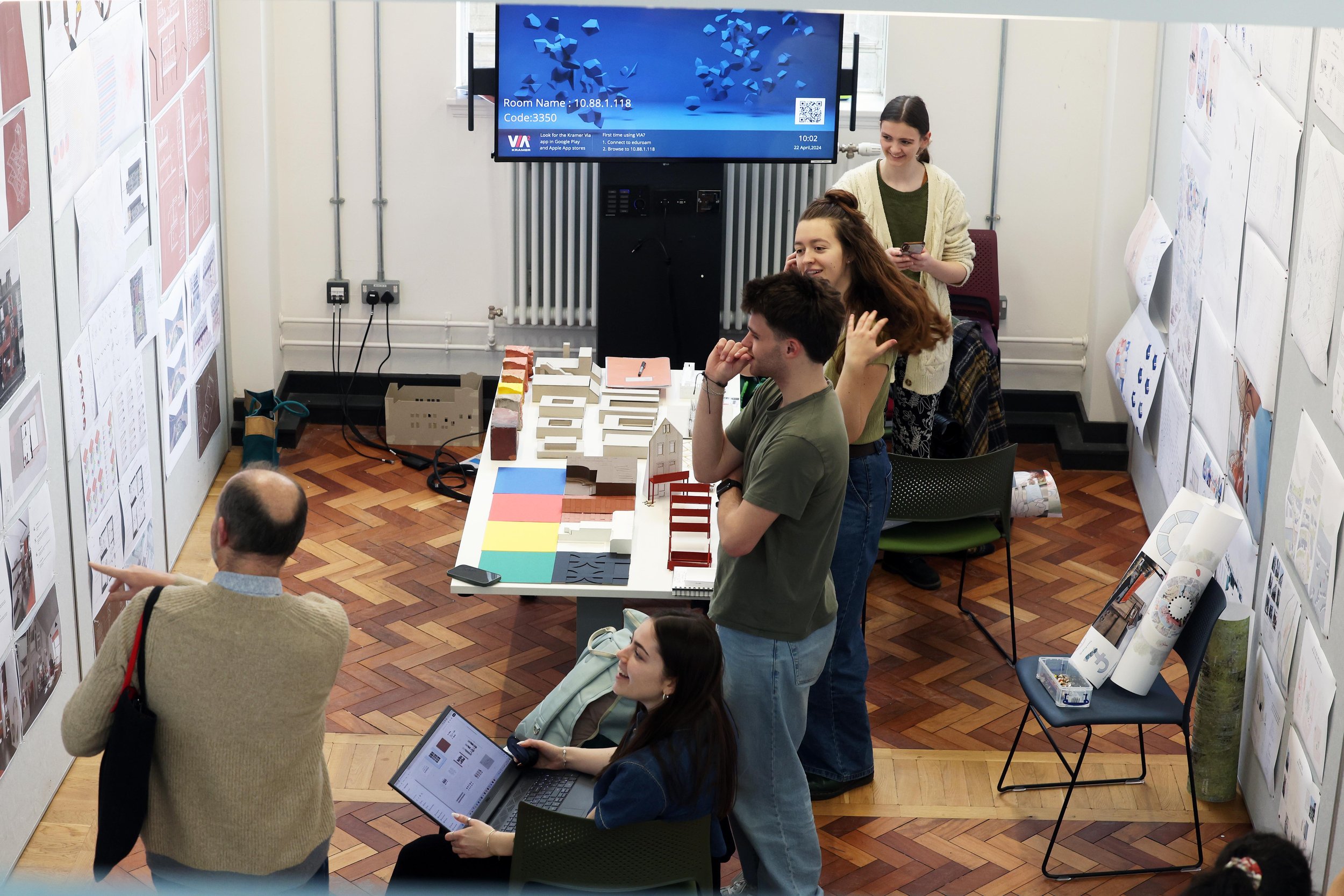 Myfyrwyr ym Mhrifysgol Caerdydd yn ystod sesiwn adborth stiwdio gyda thabl o fodelau a gwaith dylunio