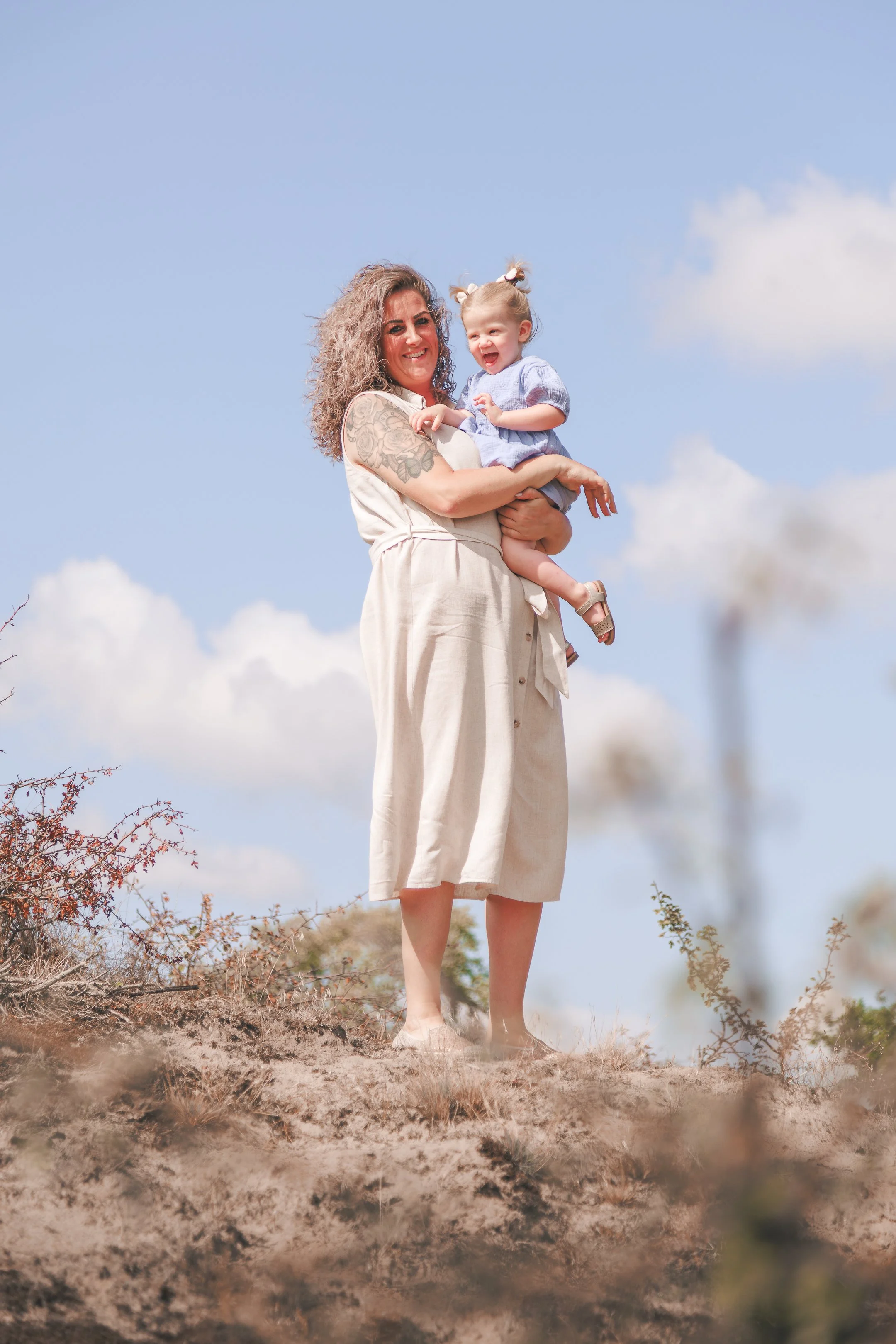 Vrouw houdt lachend een klein meisje vast terwijl ze samen buiten staan onder een heldere blauwe hemel met enkele wolken.