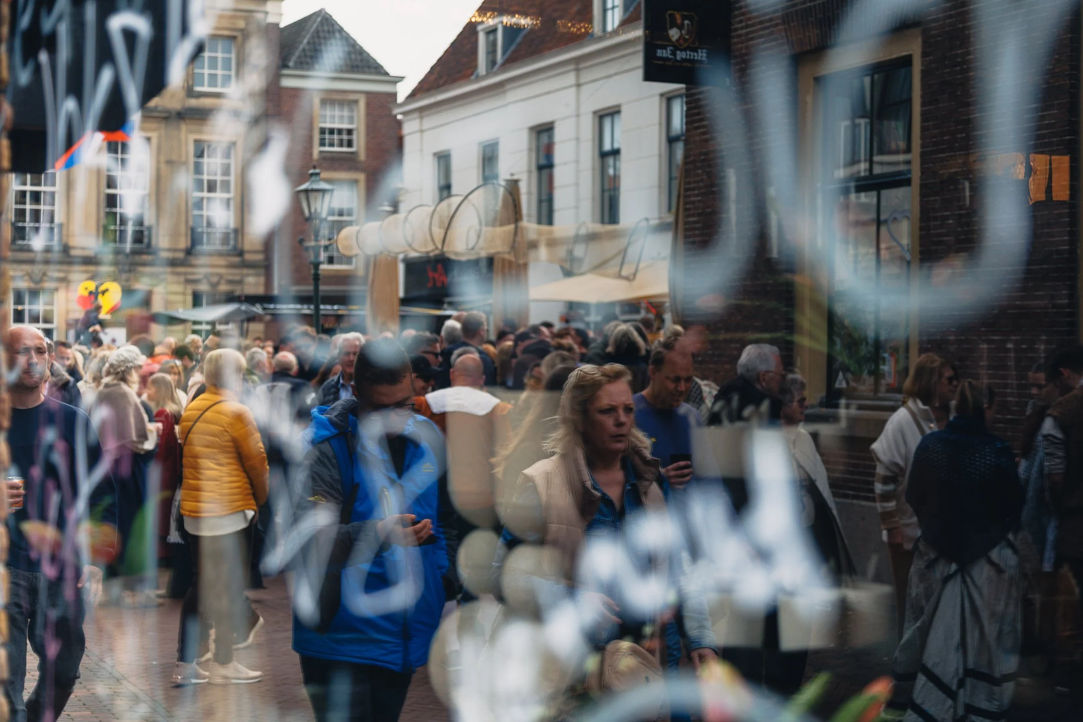 Gezichtspunten van een drukke straat in een stad, met mensen die lopen en winkeltjes op de achtergrond, gezien door een raam met een metalen hek.