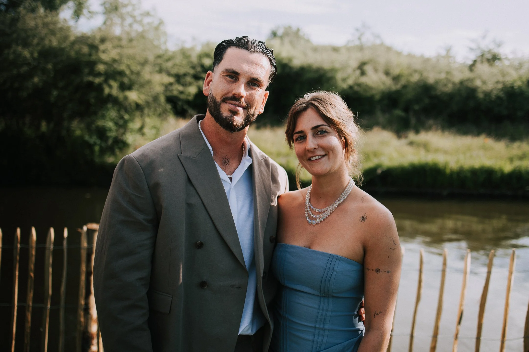Een man en een vrouw staan dicht bij elkaar buiten, met groene bomen en water op de achtergrond. Ze glimlachen naar de camera, de man draagt een grijze jas en de vrouw een blauwe jurk met sieraden.