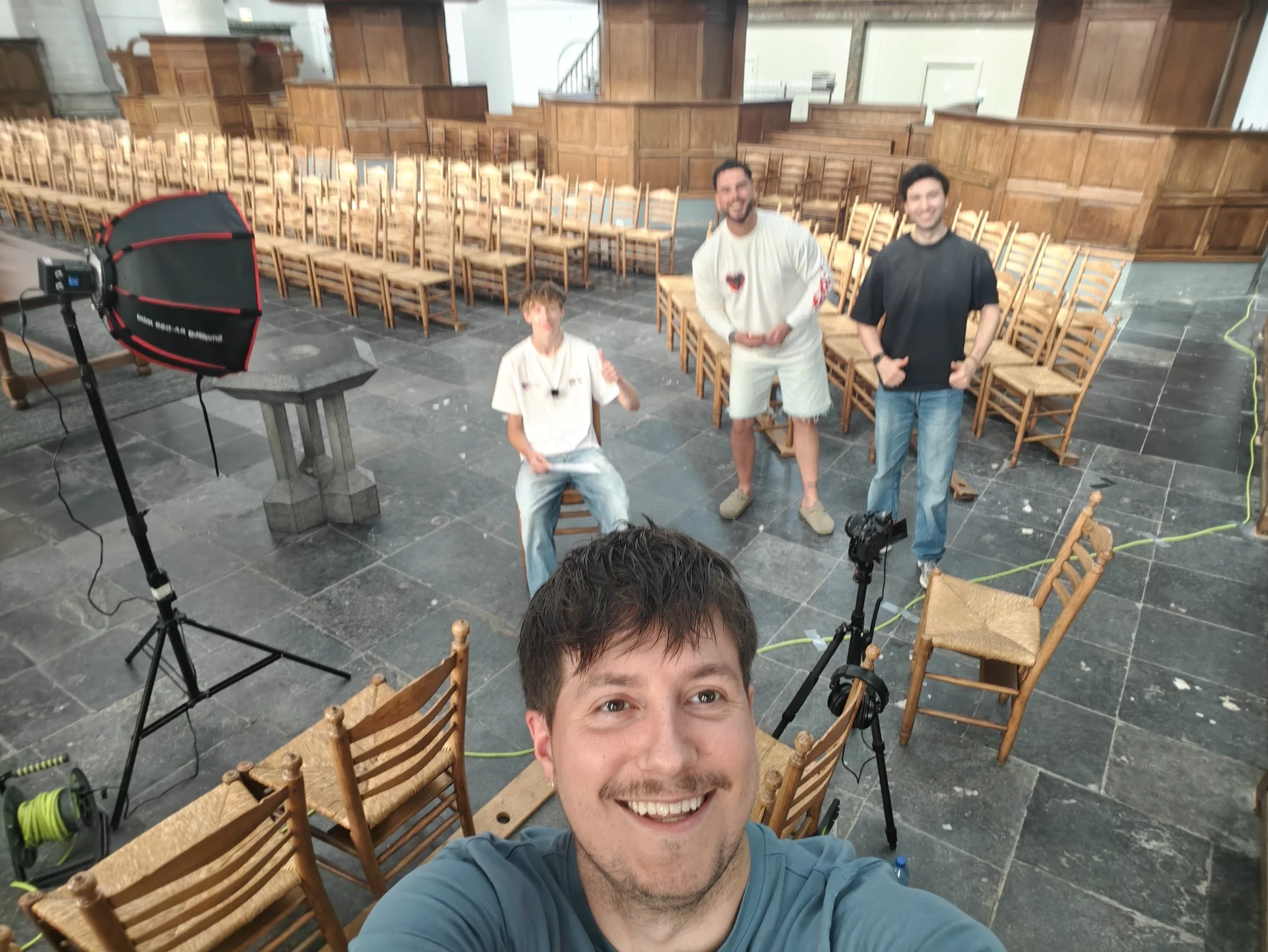 Man die een selfie neemt in een grote ruimte met houten stoelen en twee andere mannen op de achtergrond.