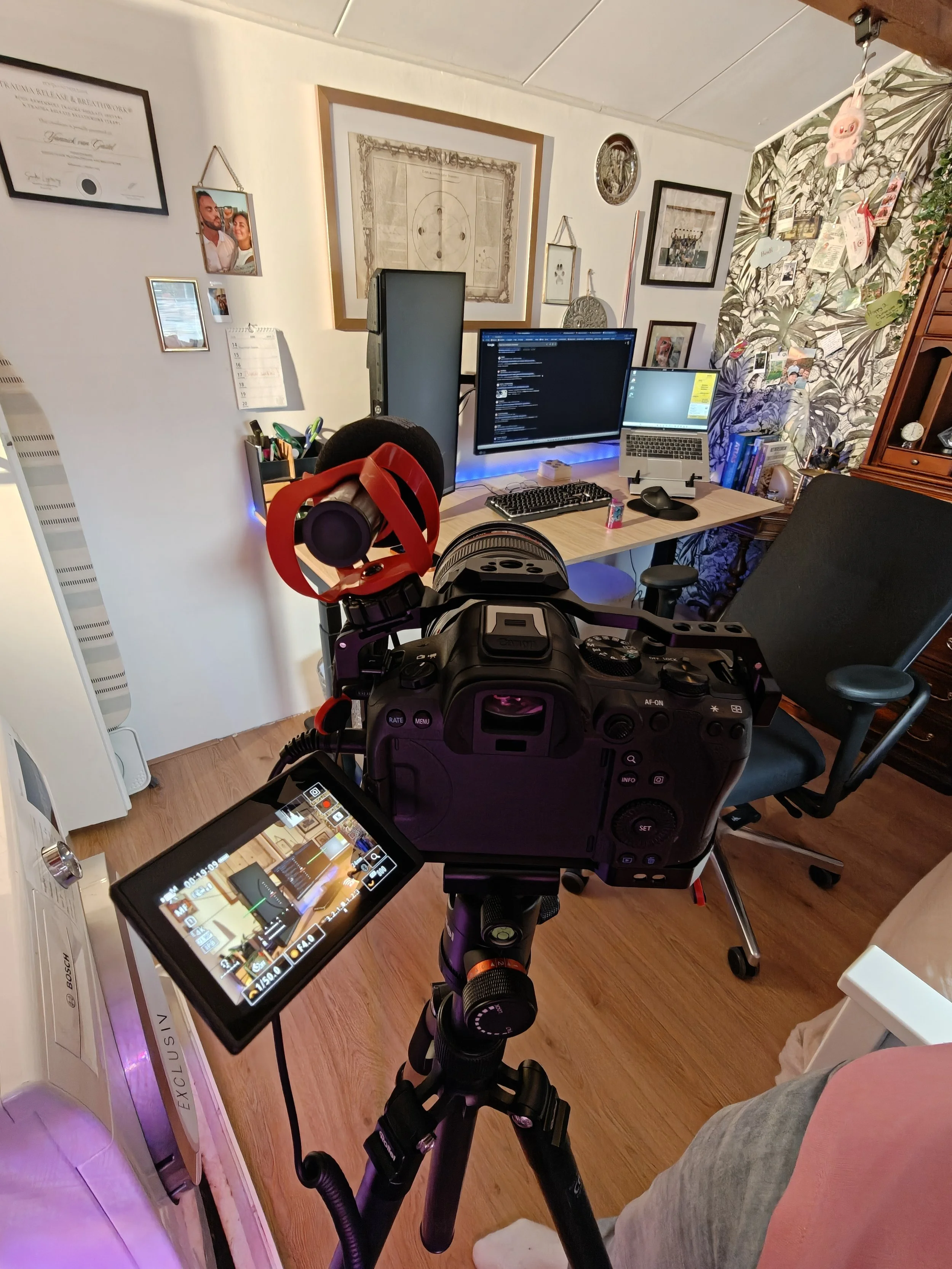 Een kamer met een bureau met twee computers, een computerfoto met videoregistratieapparatuur, en een muur met foto's en kunstwerken, inclusief een diploma en een kalender.