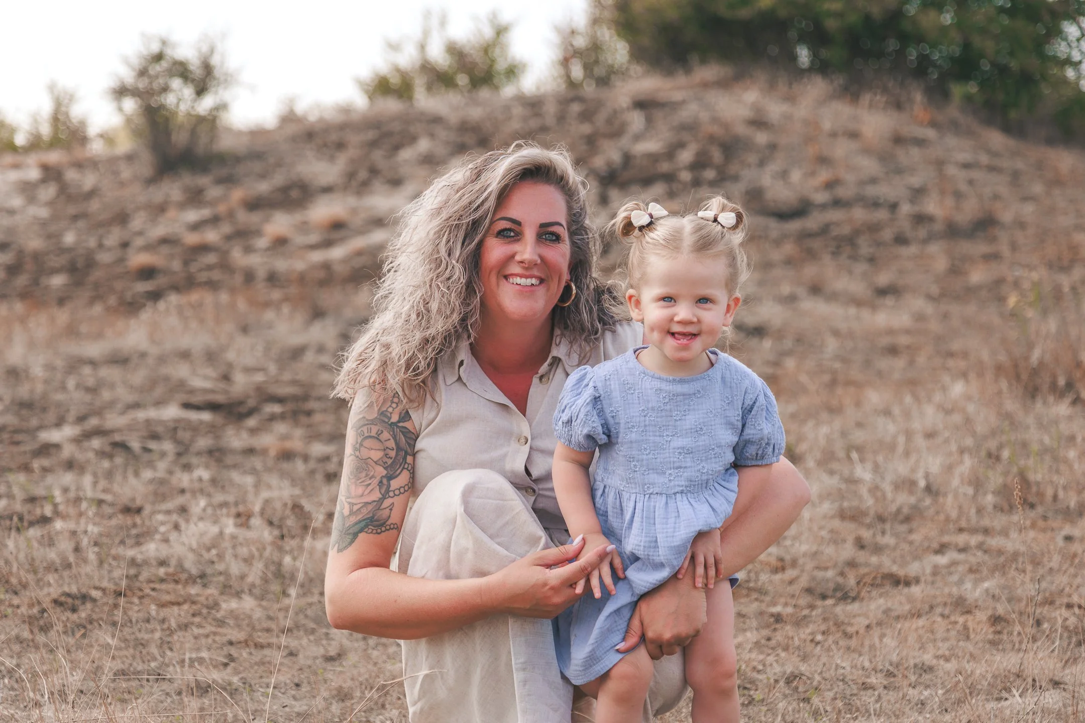 Een vrouw met lang, golvend blond haar en een tatoeage op haar arm, die een jonge meisje vasthoudt in een veld met droge planten en bomen op de achtergrond.