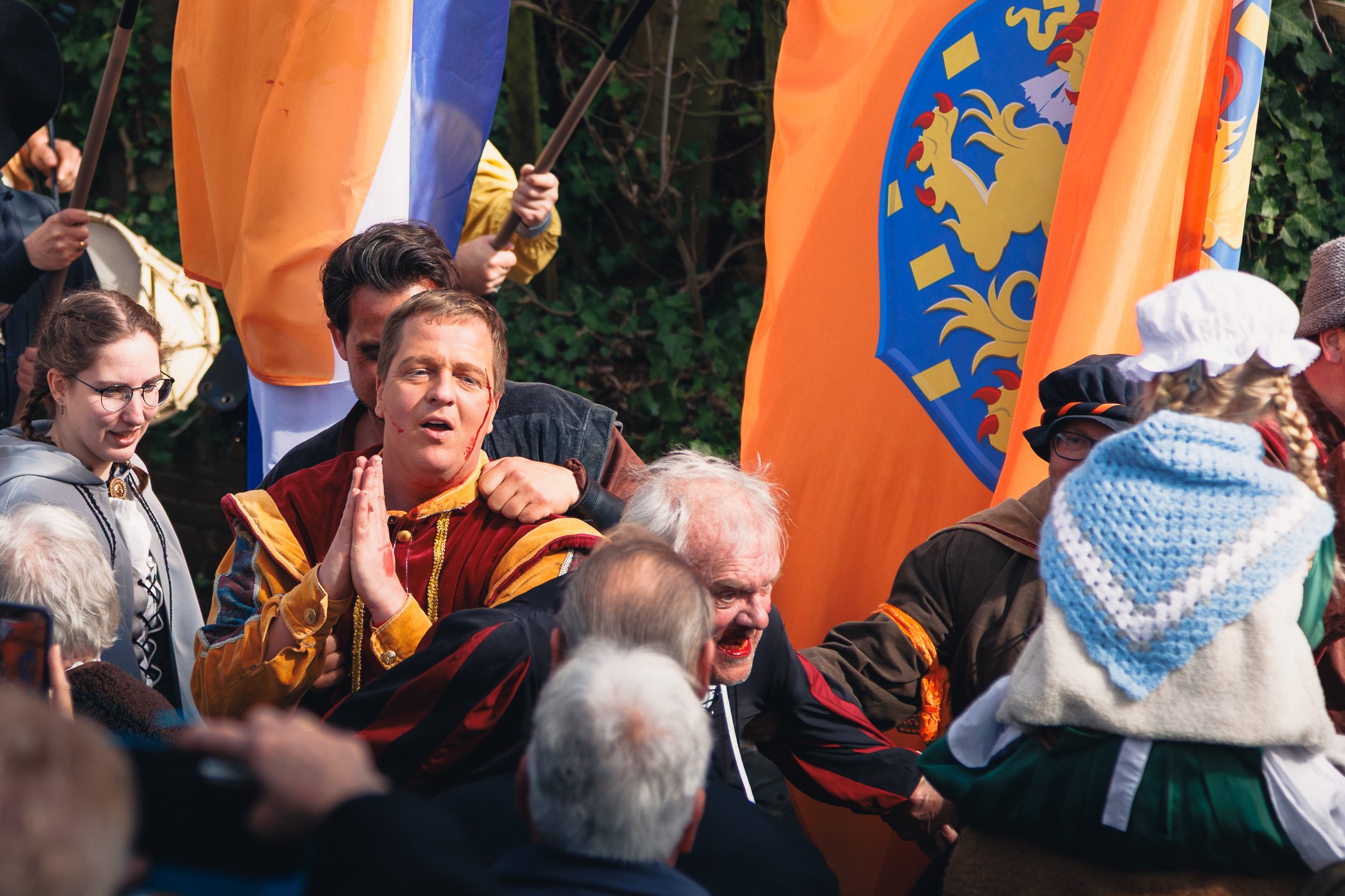 Mensen bij een kleurrijke parade of evenement, sommige mensen dragen historische of middeleeuwse kleding en vlaggen met dieren en symbolen.