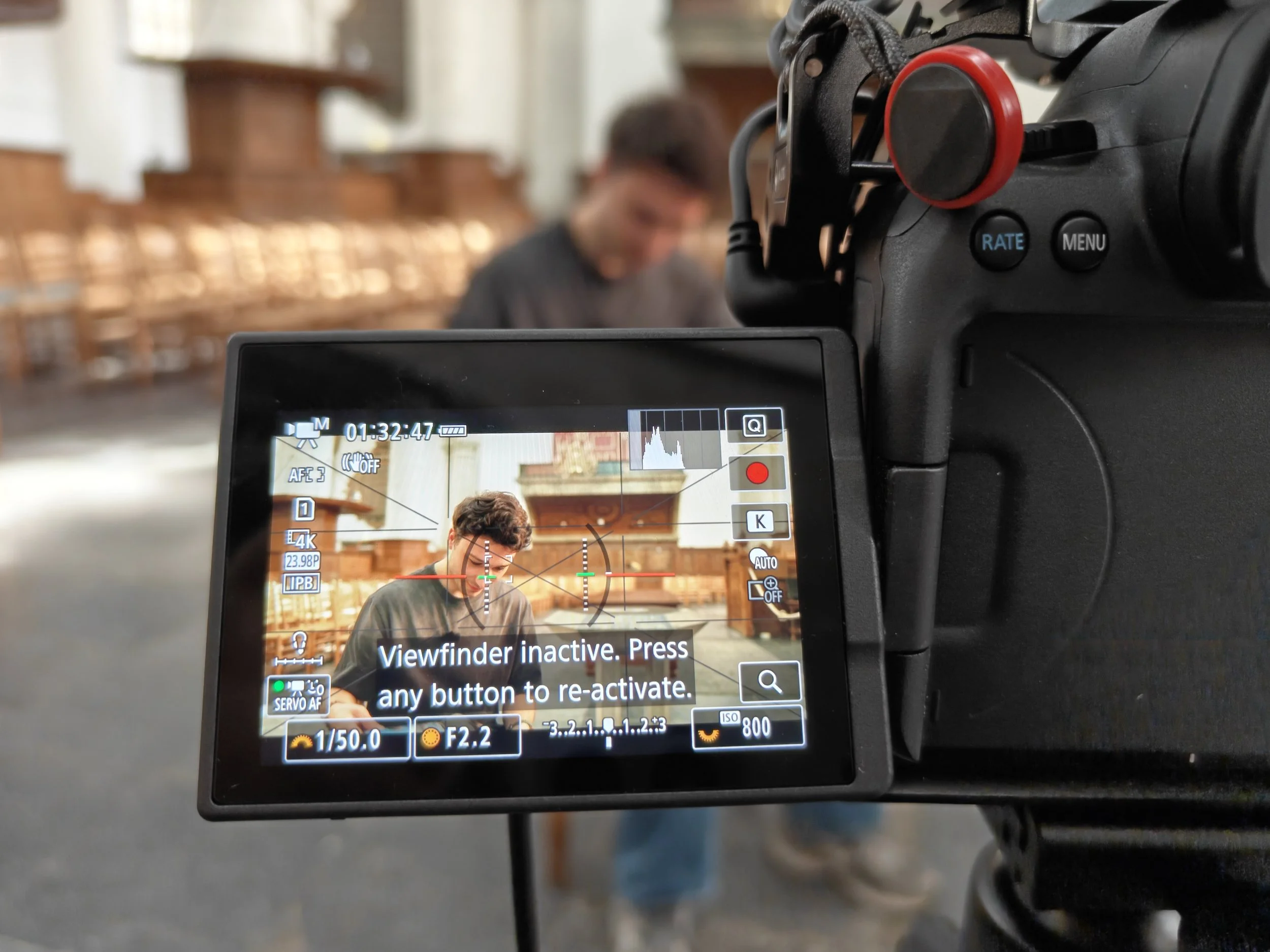 Close-up van een videocamera-instellingenmonitor waarop te zien is dat de opname is gestopt en dat de viewfinder inactief is, met een persoon die op de achtergrond te zien is en een gerestaureerde kerk.