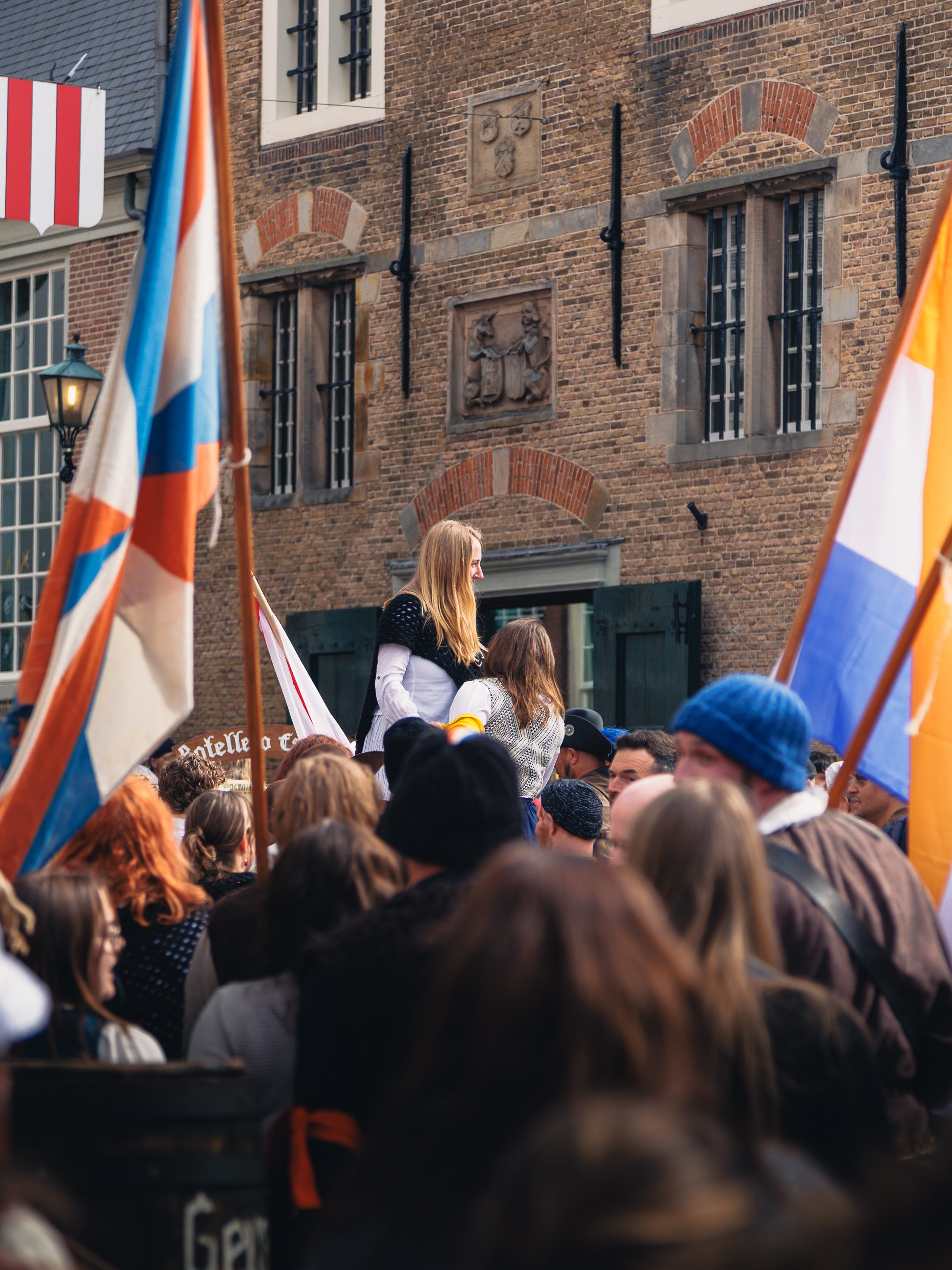 Een menigte mensen bij een buitenevenement, met vlaggen en een historische bakstenen gevel op de achtergrond.