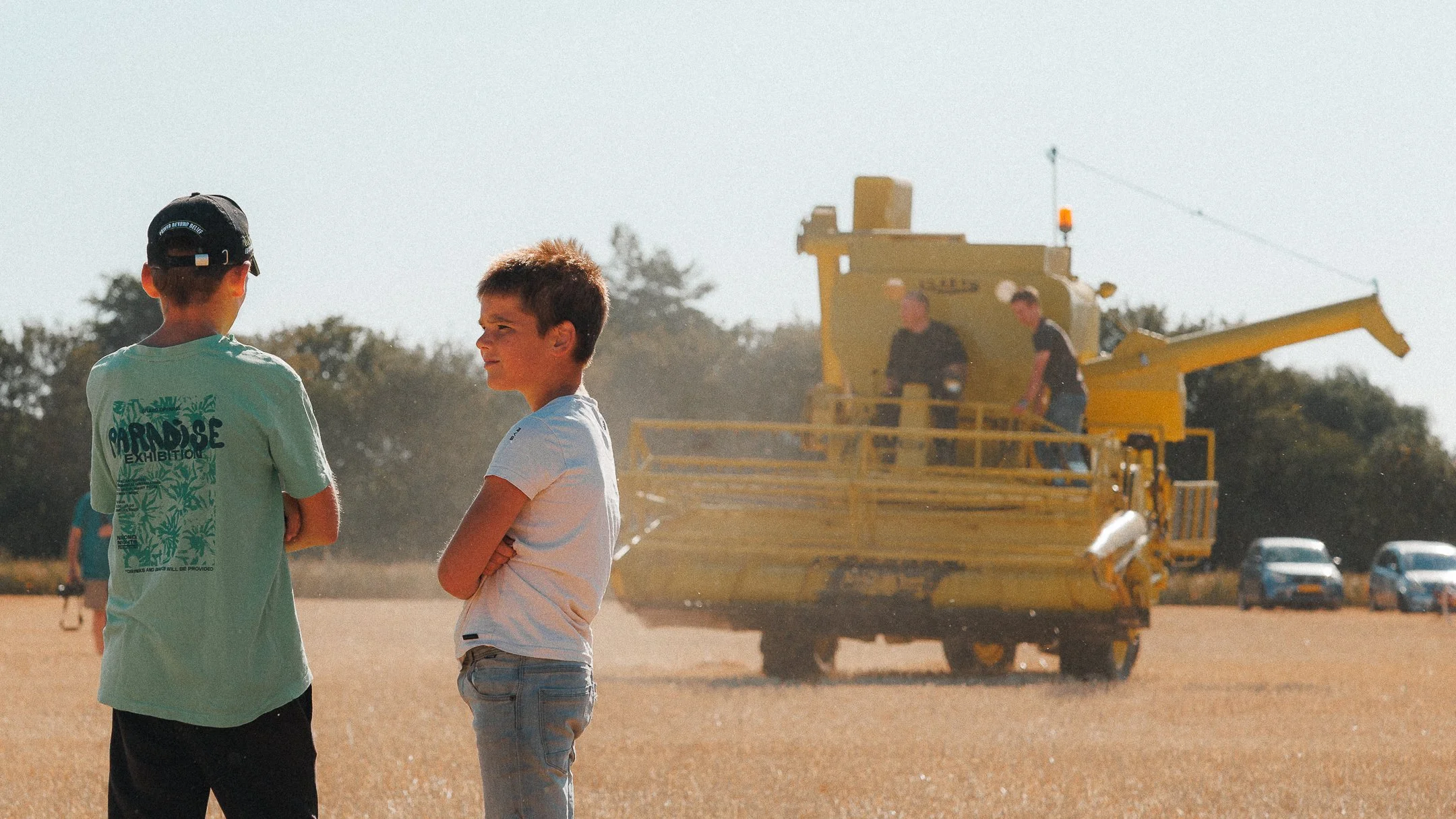 Twee jongens staan met hun armen over elkaar op een veld, terwijl een zandwagen en auto's op de achtergrond te zien zijn op een zonnige dag.
