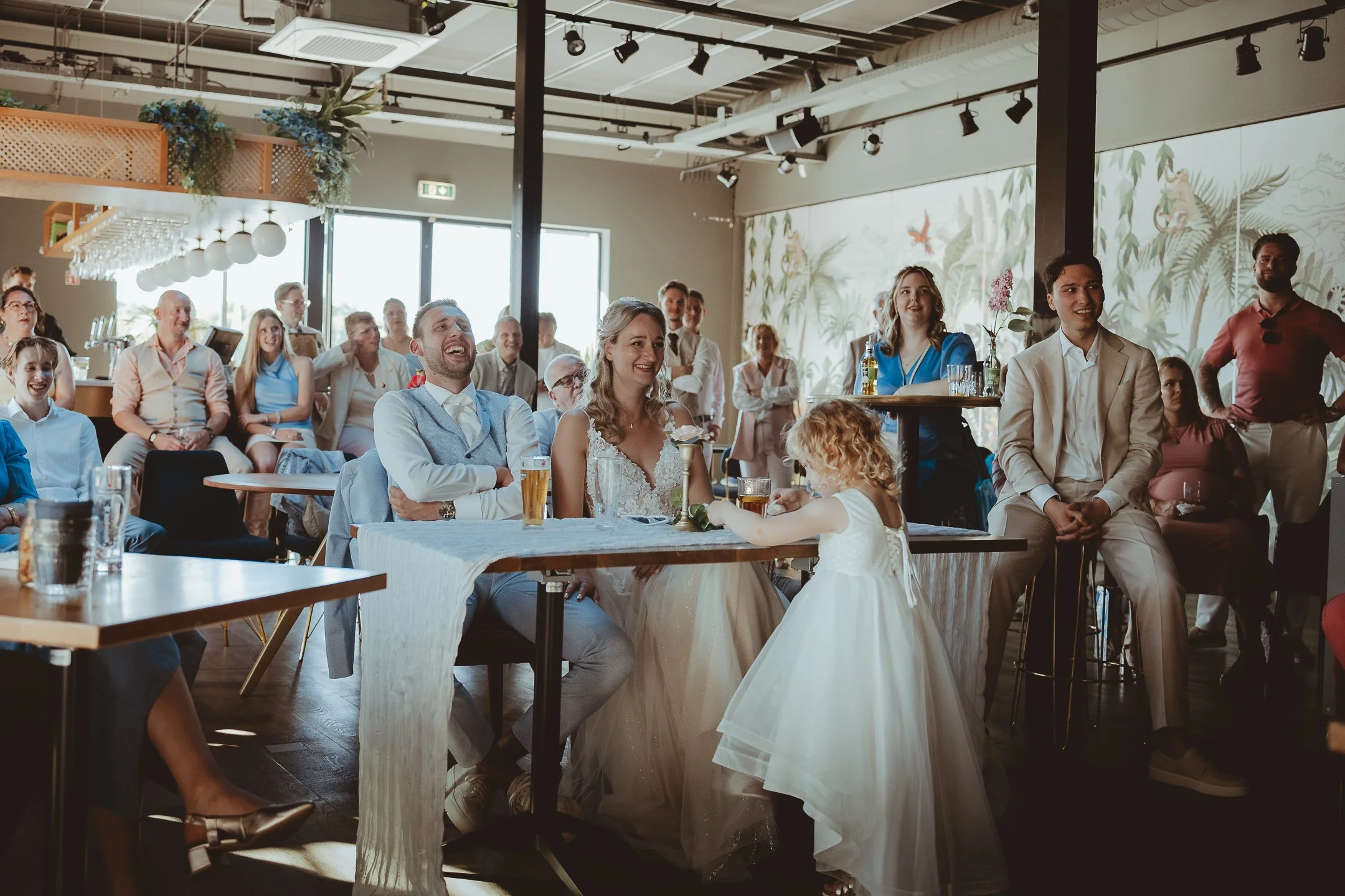 Een groep mensen zit en staat in een feestzaal, lachend en kijkend naar een optreden of spreker. Er zijn twee meisjes in witte jurken vooraan, mogelijk bruidjes. De sfeer is vrolijk en feestelijk met decoraties en grote ramen die natuurlijk licht bin