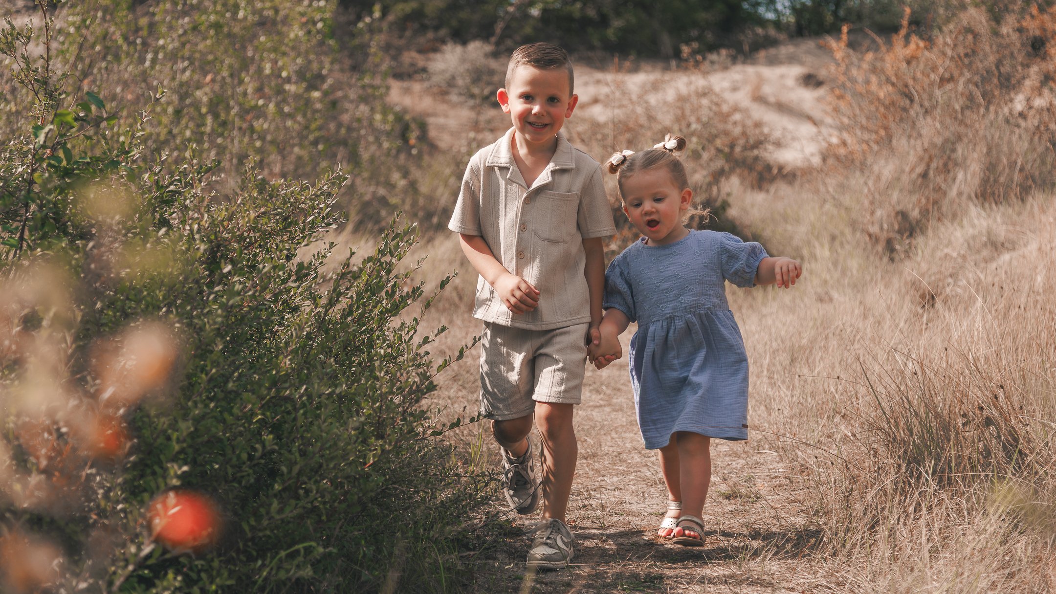 Twee jonge kinderen, een jongen en een meisje, wandelen hand in hand door een veld met dorre grashalmen en struiken, omgeven door een natuurlijke omgeving met struiken en bomen in de achtergrond.