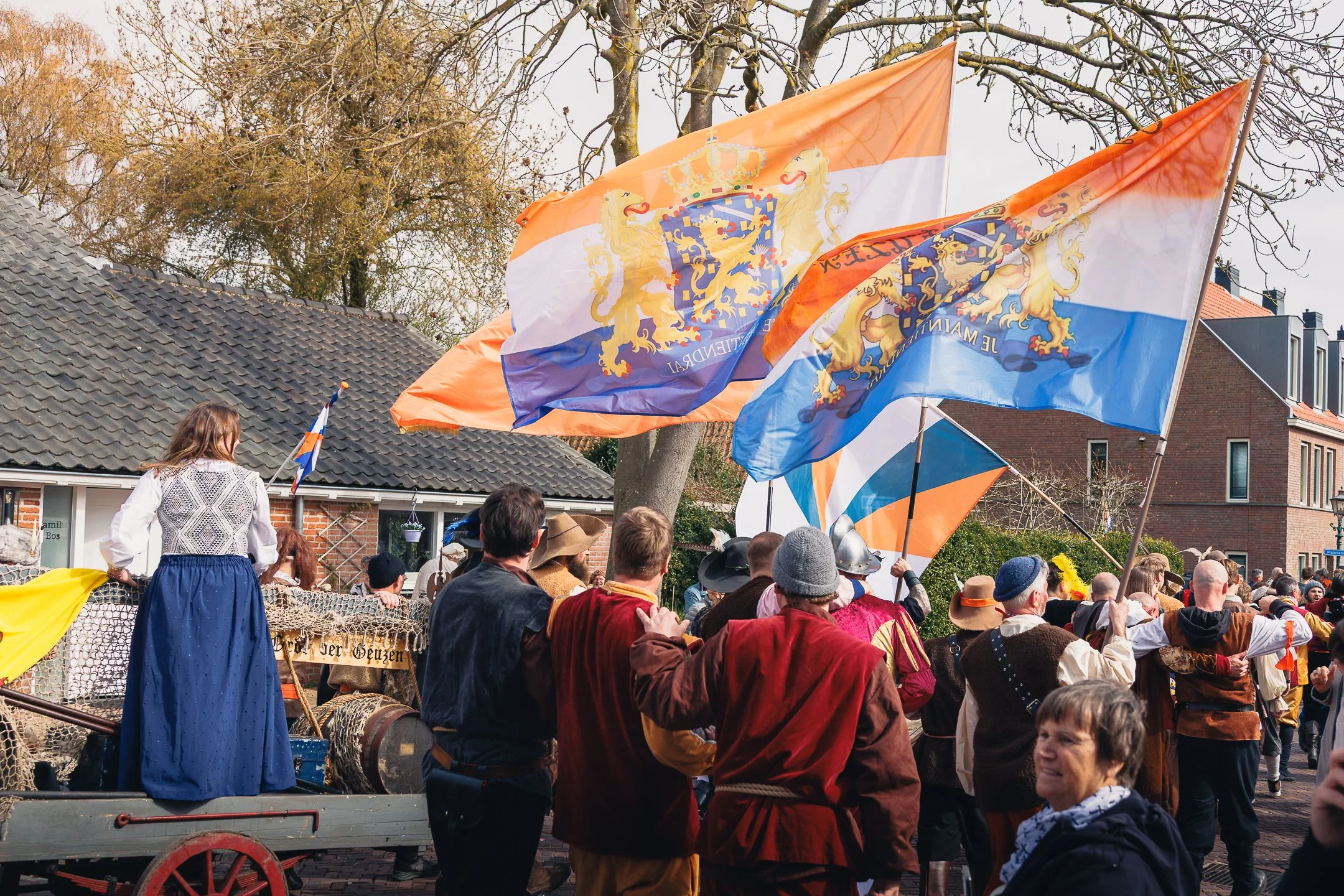 Een groep mensen in historische kostuums die deelnemen aan een optocht of viering met Nederlandse vlaggen en leeuwensymbolen, omringd door huizen en bomen.