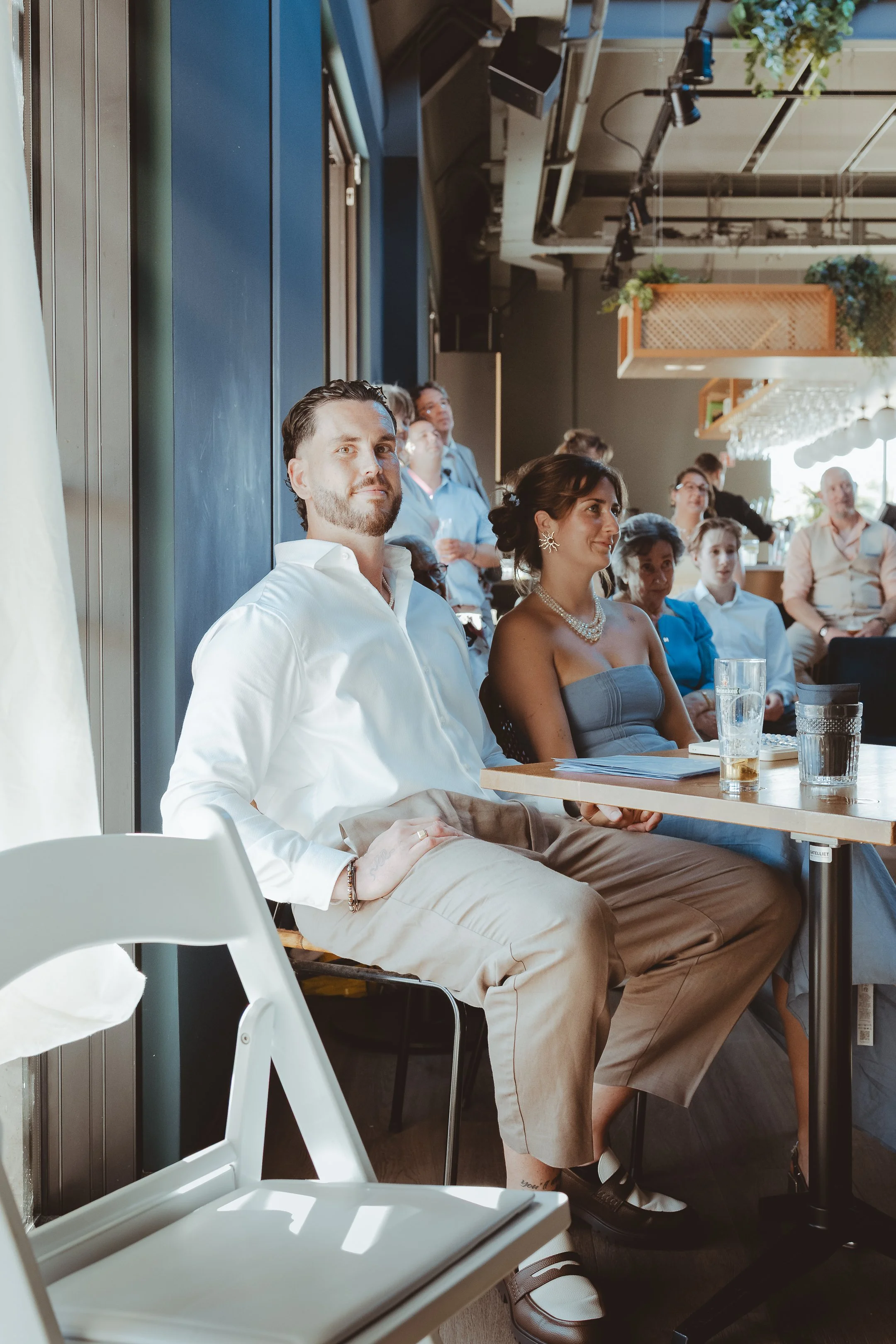 Binnen in een modern café of restaurant, mensen zitten aan tafels en luisteren naar een spreek- of voordracht. Twee mensen op de voorgrond, een man met een witte button-down shirt en beige broek, een vrouw met een donkerblauwe jurk en sieraden.