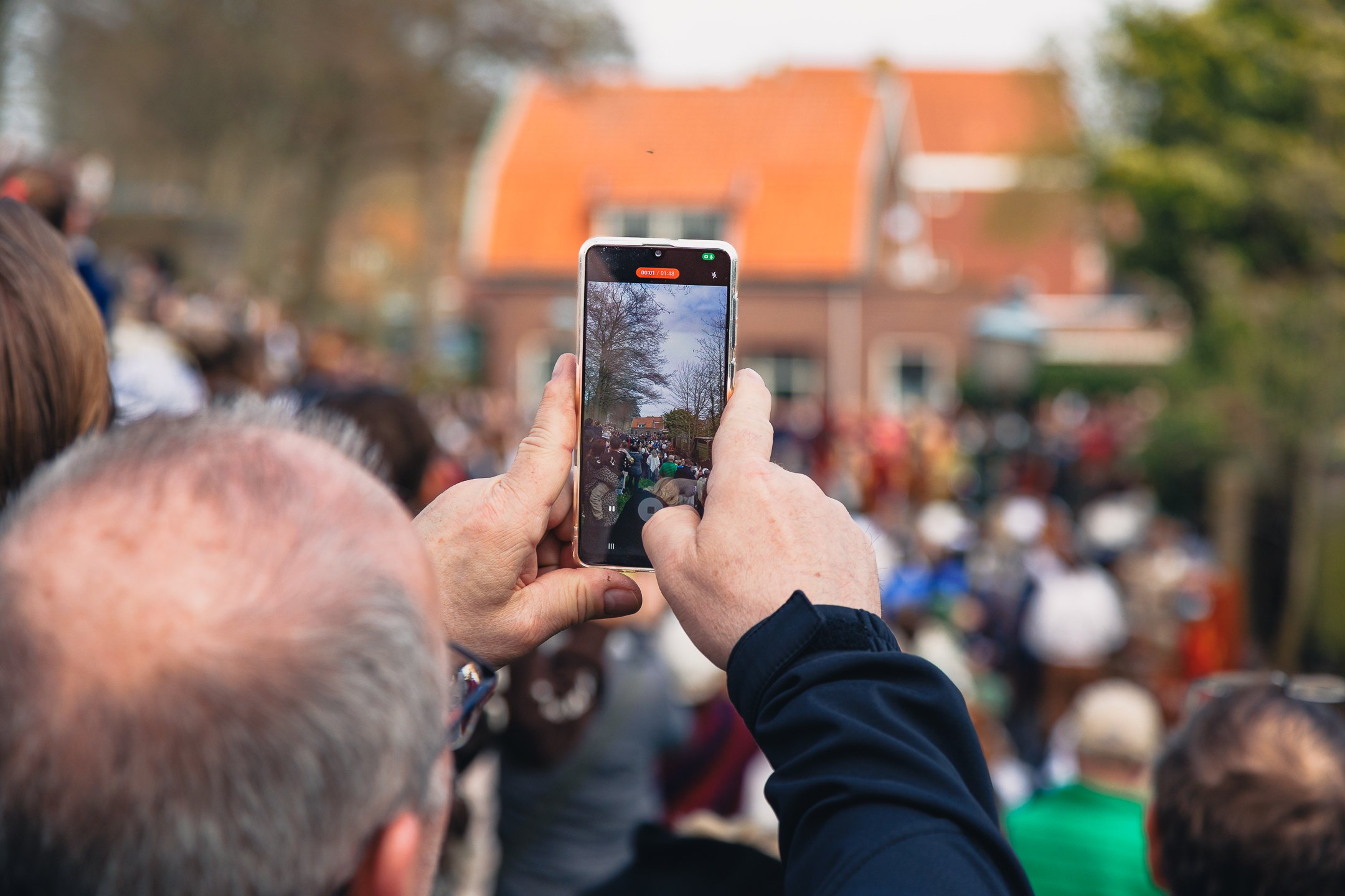 Een persoon houdt een smartphone omhoog om een groep mensen vast te leggen die bij een evenement in een straat staan, met een huis met een oranje dak op de achtergrond.