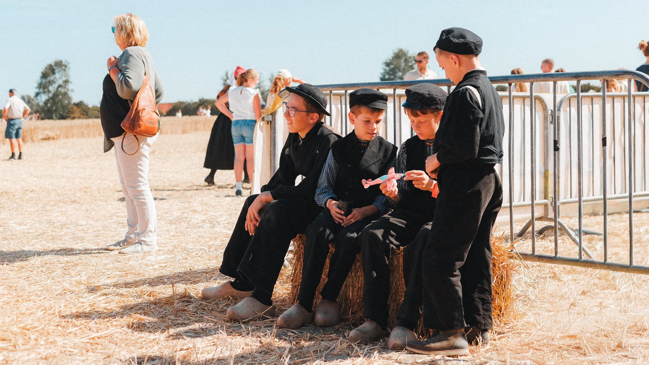 Vier kinderen in zwarte traditionele kleding en hoeden zitten op stro in een open veld, met een metalen afscheiding erachter, terwijl andere mensen in de achtergrond wandelen en staan onder een heldere blauwe hemel.